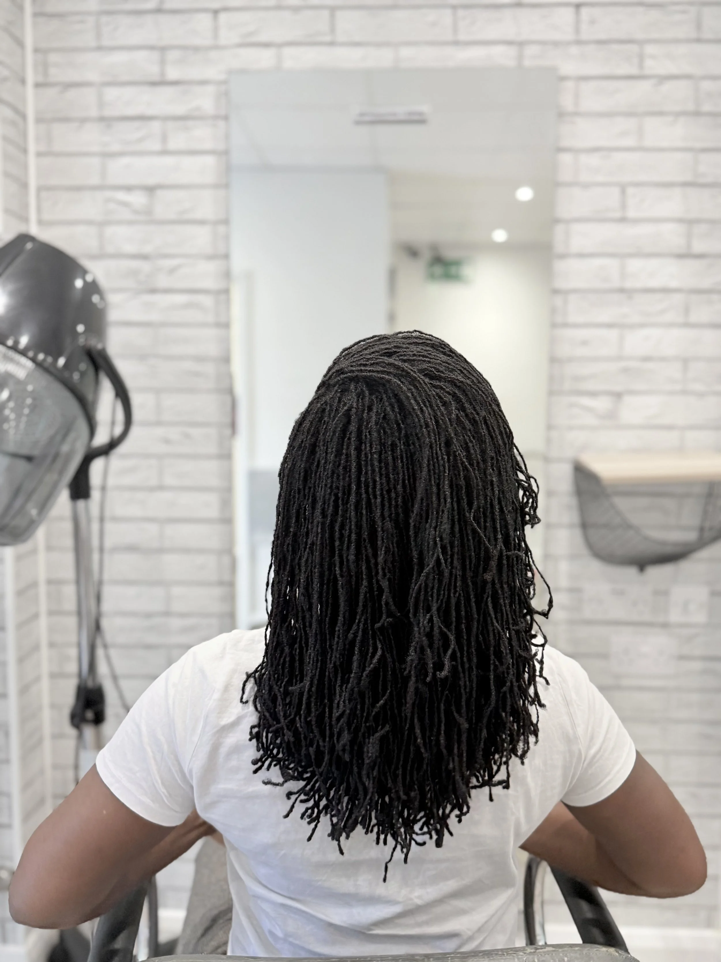 Person with sisterlocks sitting with arms behind chair, seen from behind, in front of a mirror and hairdryer at a salon or barbershop.