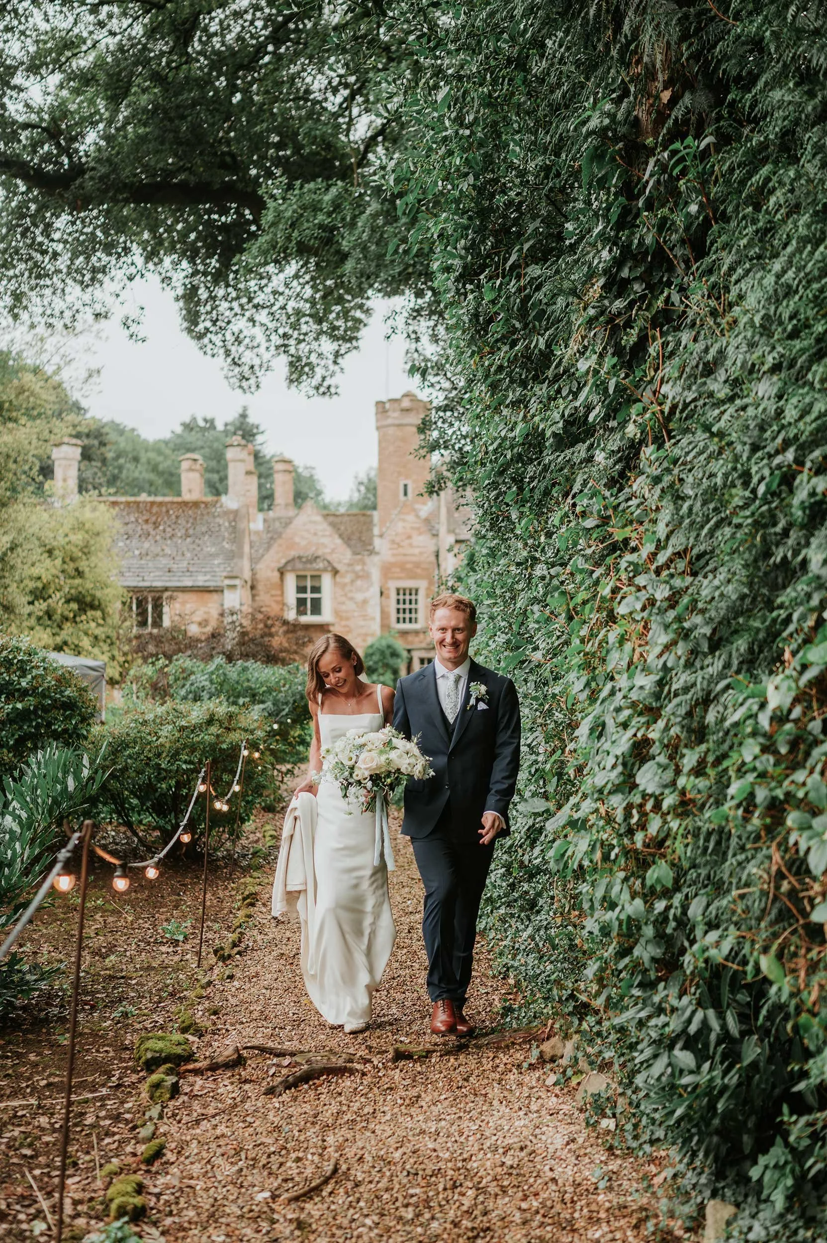 A bride and groom walking along a garden path, smiling, with Bisbrooke Hall in the background, trees overhead, and string lights on the side.