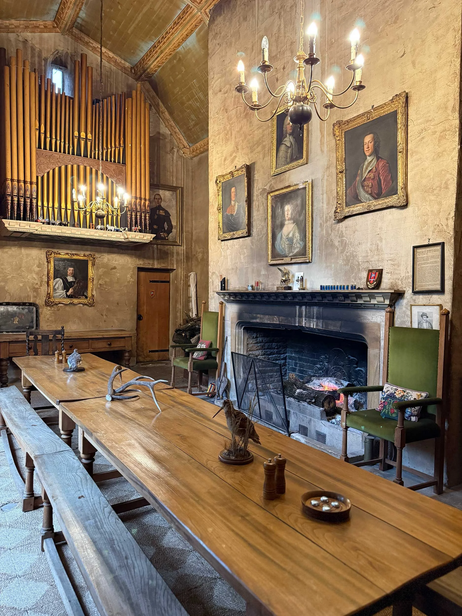 A cozy living room with a large wooden dining table, green chairs, and a decorative fireplace with a fire burning. The wall is adorned with portraits, a chandelier hangs from the ceiling, and an organ is visible in the corner.