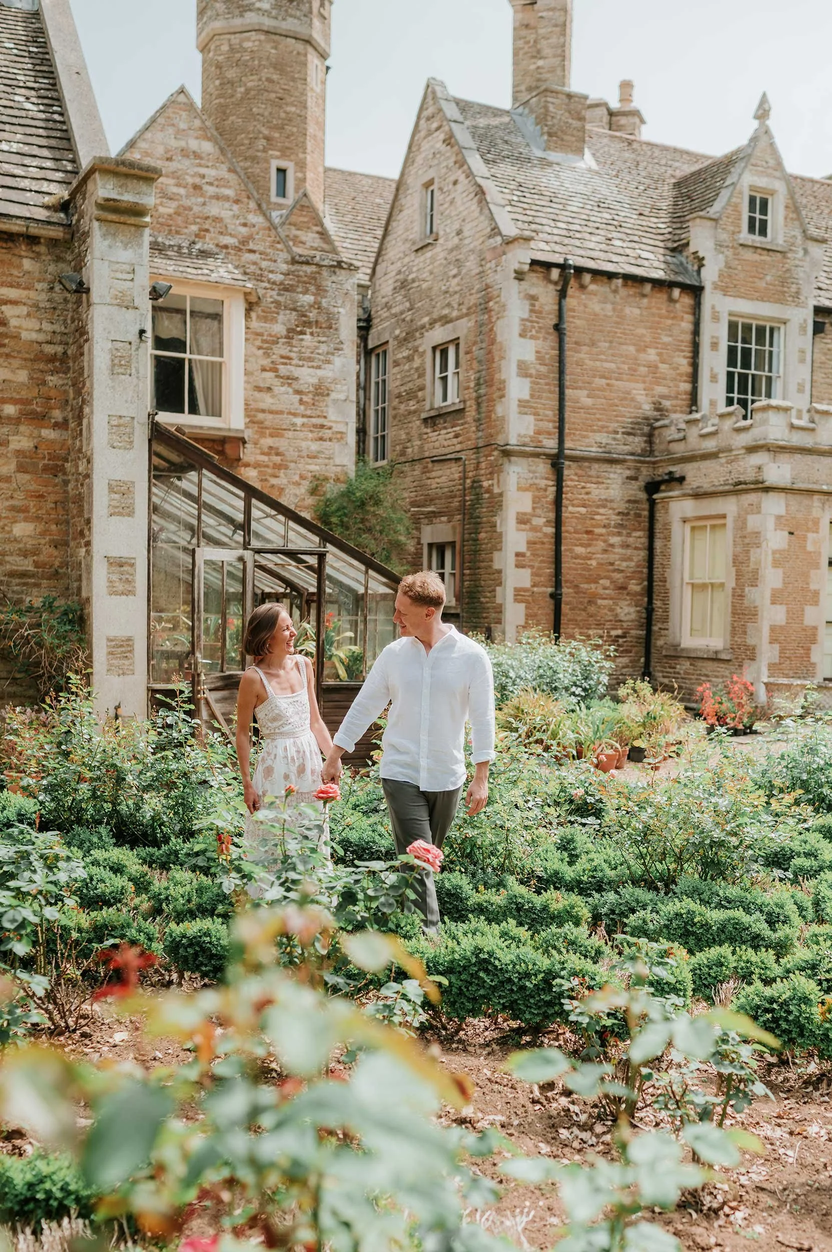 A young couple holding hands and walking through a lush garden outside a historic brick building with turrets and large windows.