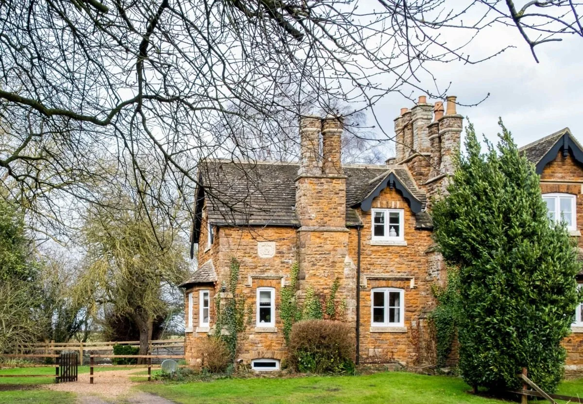 A historic brick cottage with multiple chimneys, white-framed windows, and black decorative trim, surrounded by trees and a well-maintained lawn.