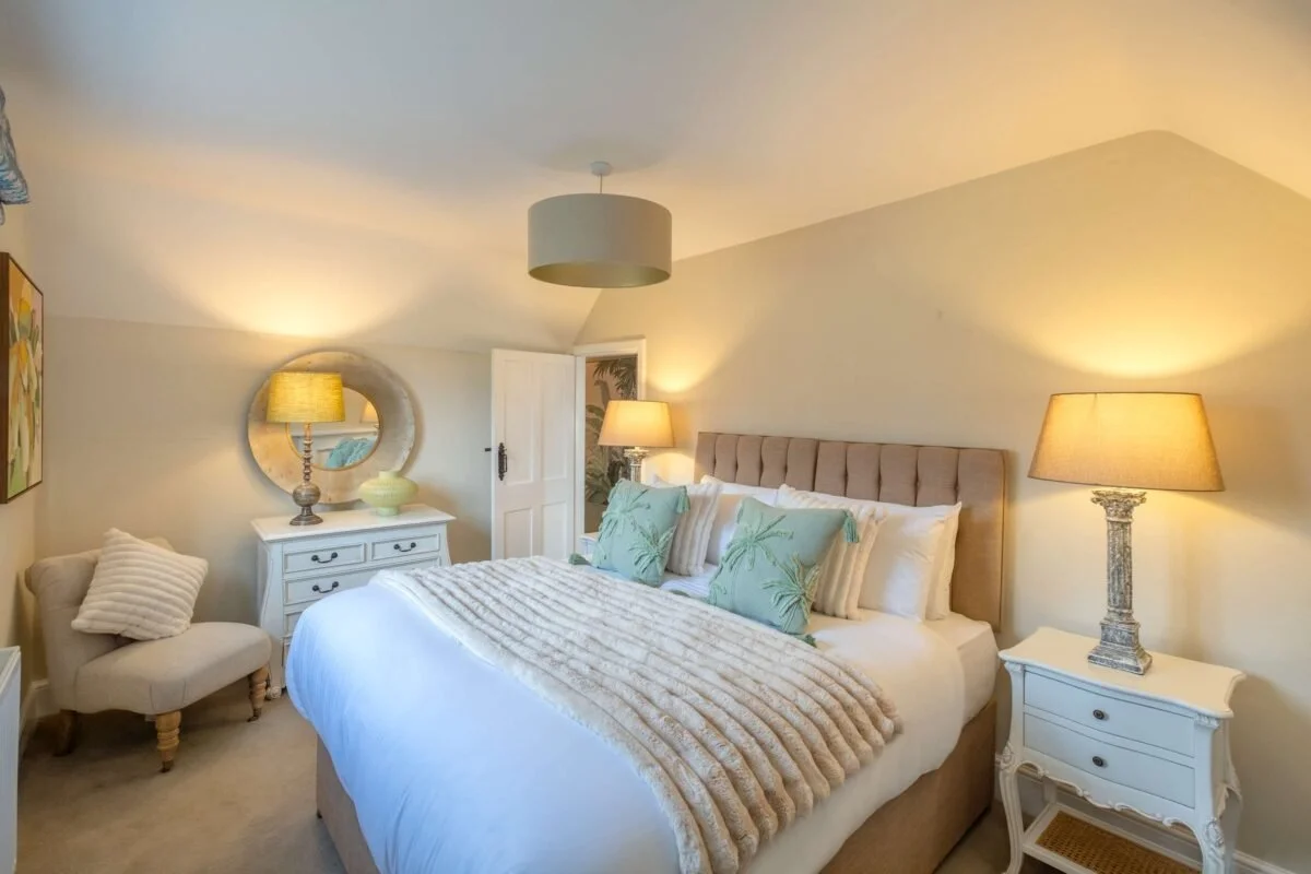 A cozy bedroom with a large bed featuring a beige upholstered headboard, layered with pillows and a soft beige throw blanket. Beside the bed is a white nightstand with a decorative lamp. To the left, there is a small upholstered chair with a striped pillow, a white dresser with a round mirror and a lamp, and a door in the background. The room has soft lighting from multiple lamps, creating a warm and inviting atmosphere.