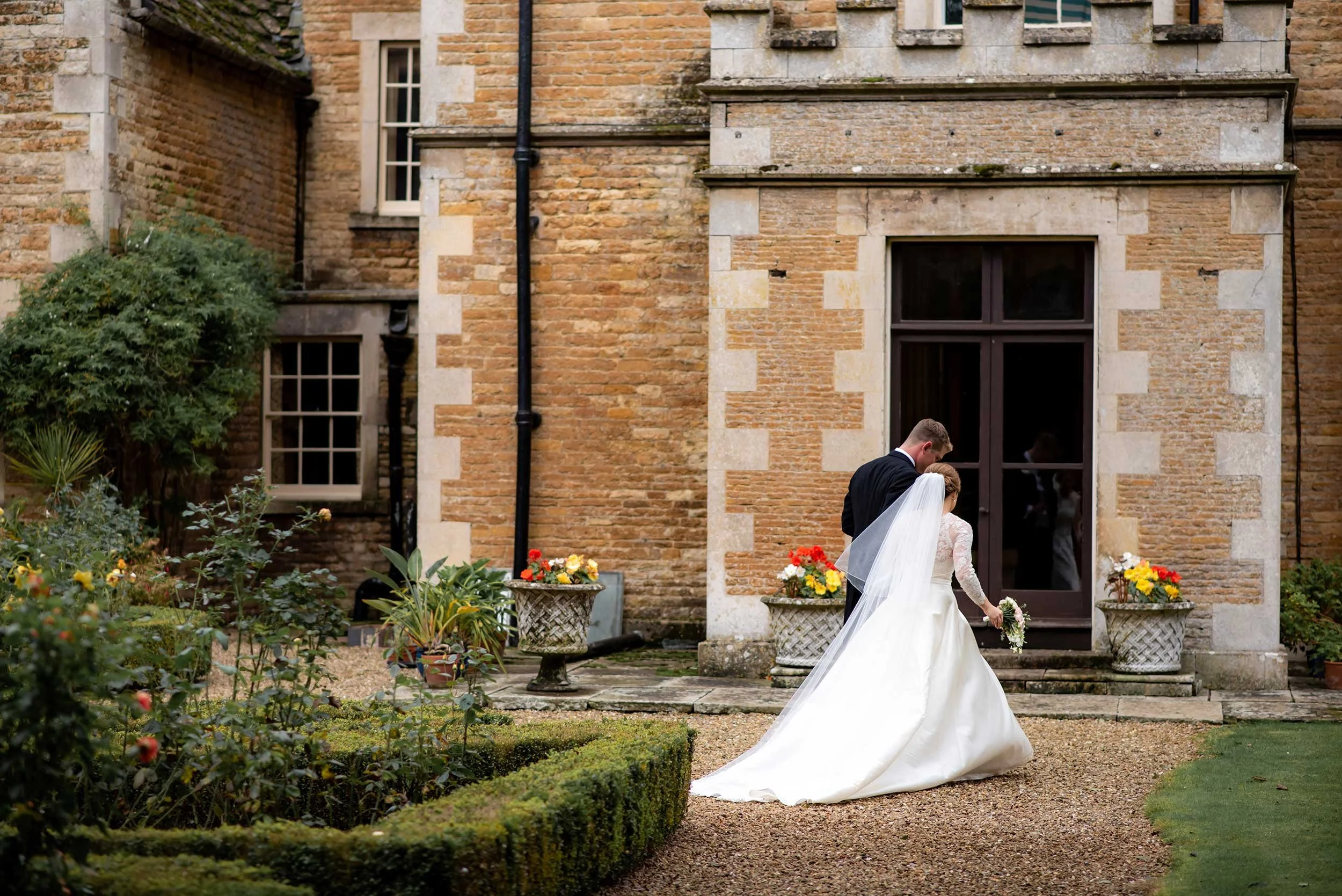 A bride and groom walking together outside a brick building, with the bride in a white wedding dress and veil holding a bouquet, and the groom in a black suit, near planters with colorful flowers.