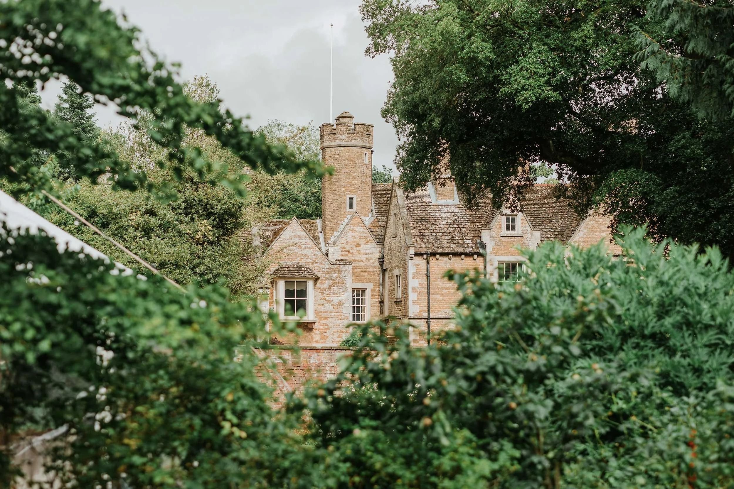 Bisbrooke Hall manor house partially obscured by dense green foliage and trees, with a cloudy sky in the background.