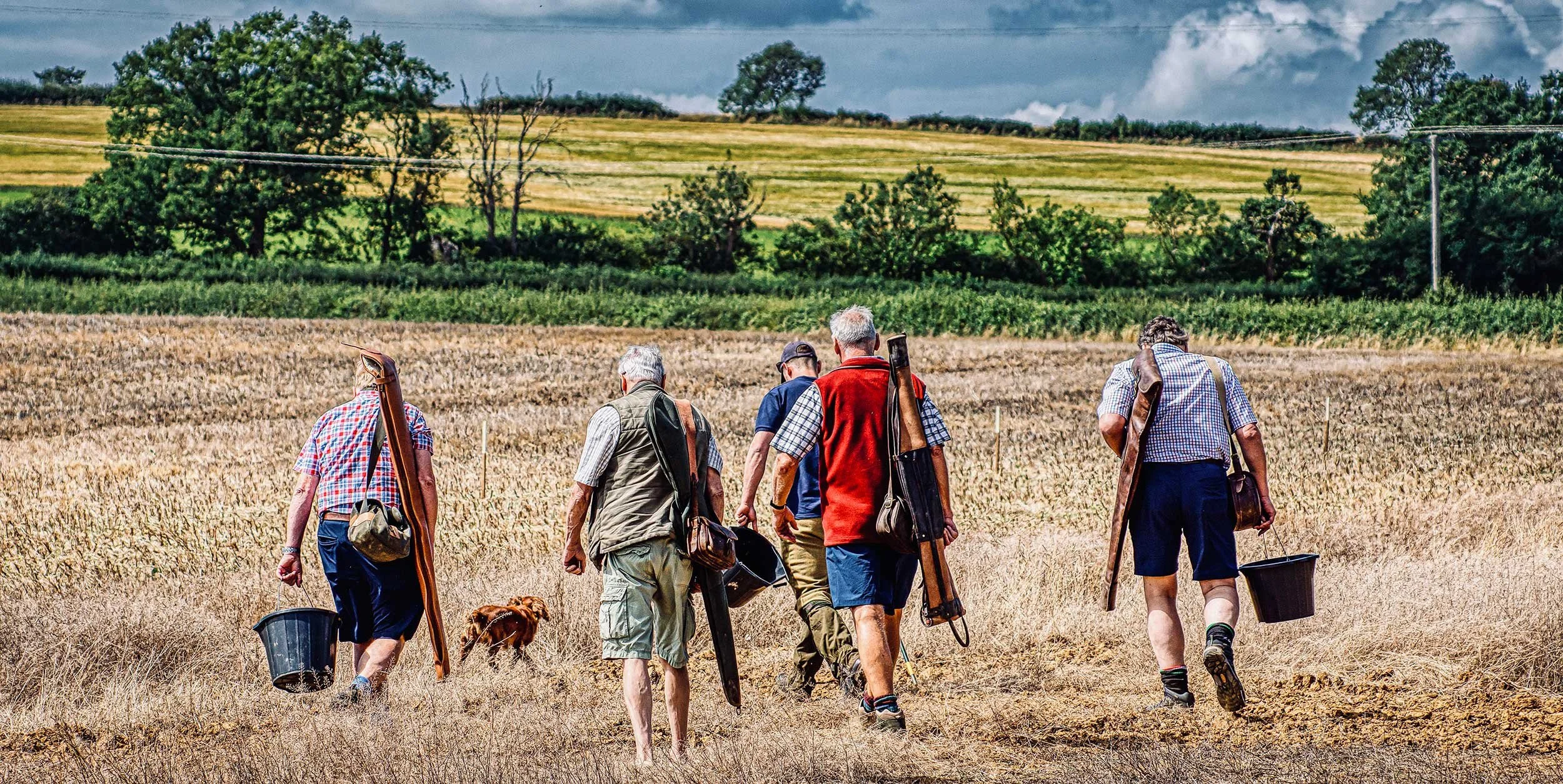 Six men walking through a field with hunting gear and a dog, green trees and hills in the background.