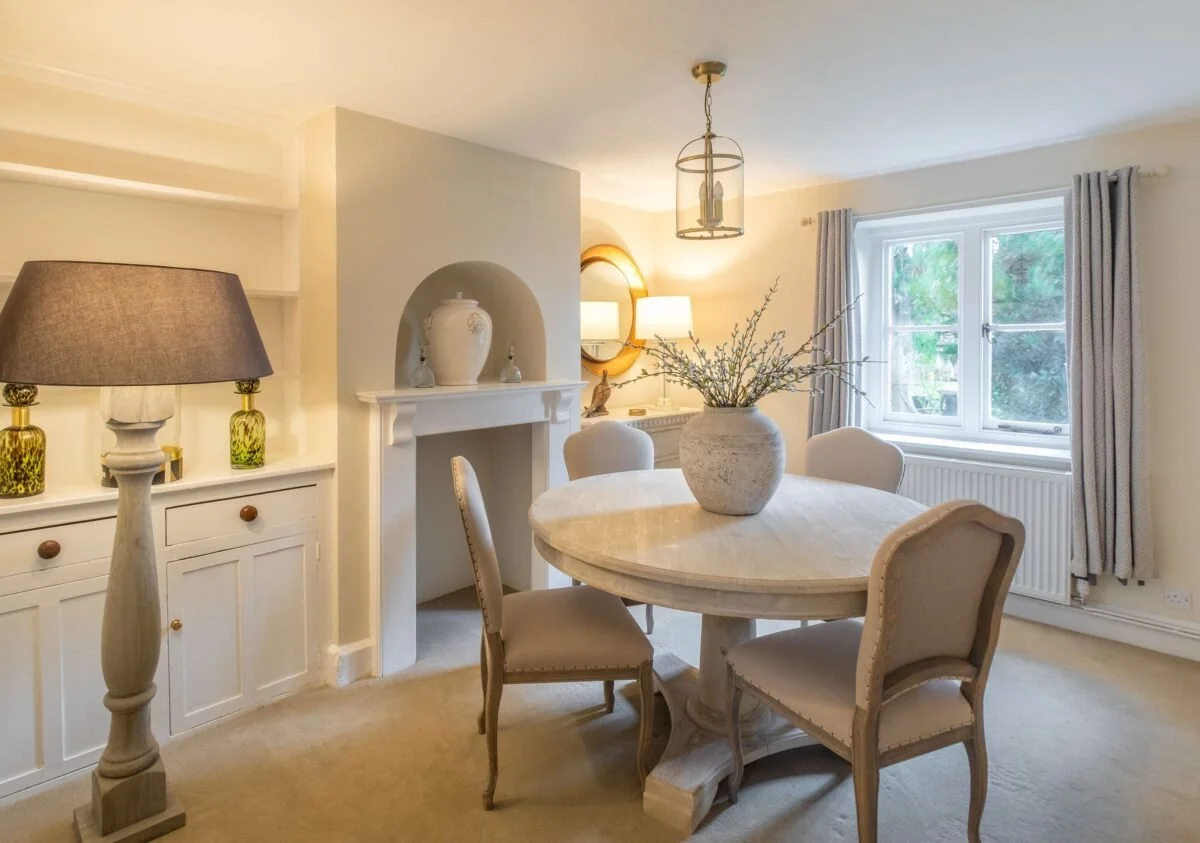 Elegant dining room with a round table, four upholstered chairs, a large vase with branches, a fireplace with decorative vases, and a window with curtains.