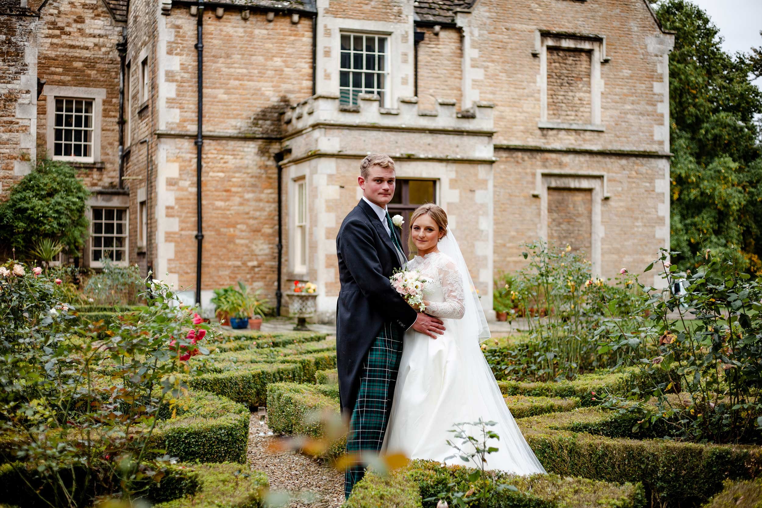 A bride and groom standing in a garden in front of Bisbrooke Hall, celebrating their wedding. The groom wears a black coat and tartan kilt, and the bride wears a long white wedding dress and holds a bouquet of flowers.