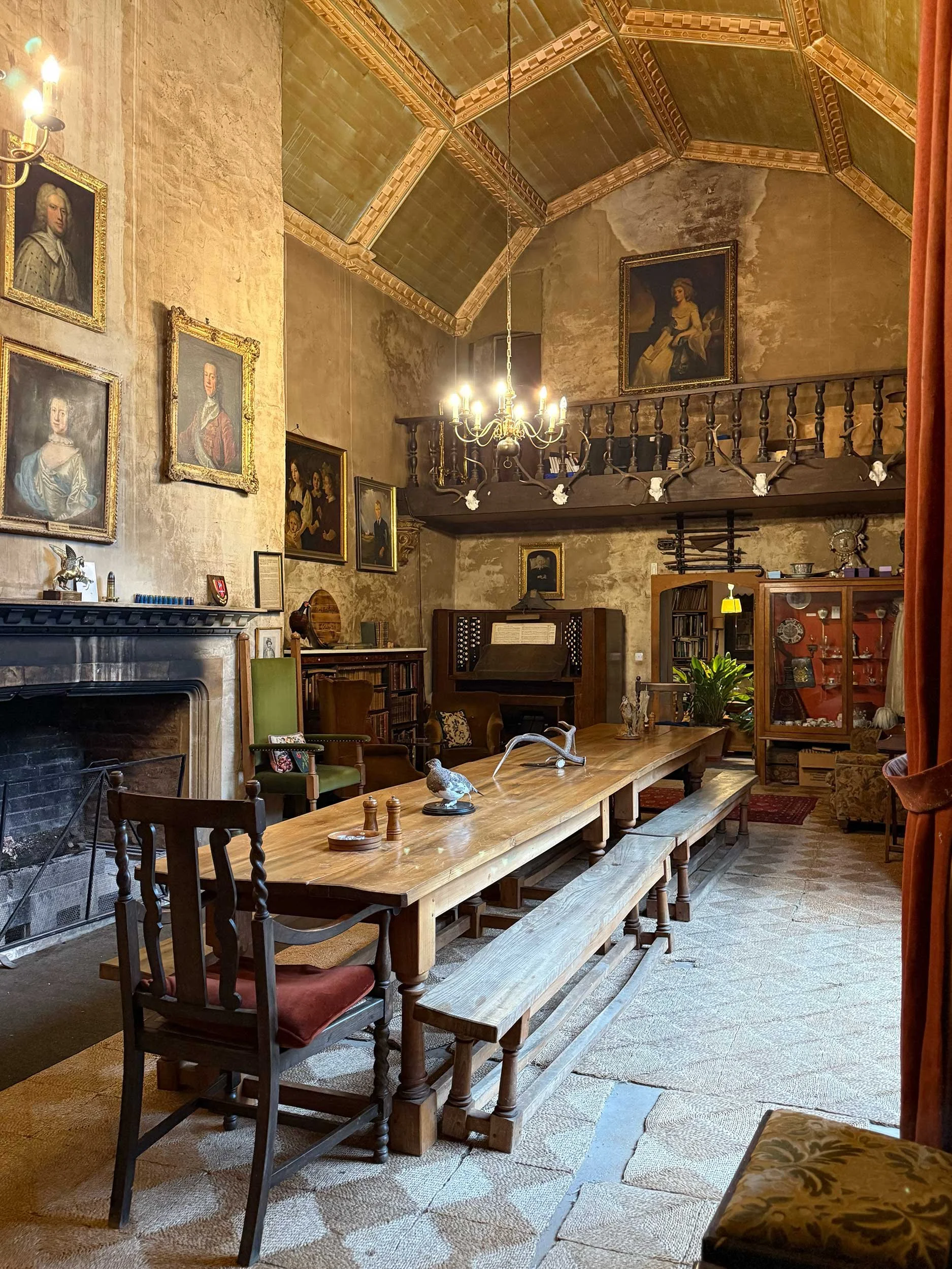 A historic dining room with vintage furniture, including a long wooden table, chairs, and a bench. The room has aged textured walls, oil portraits hanging on the walls, a chandelier, and a fireplace, creating an antique, elegant atmosphere.