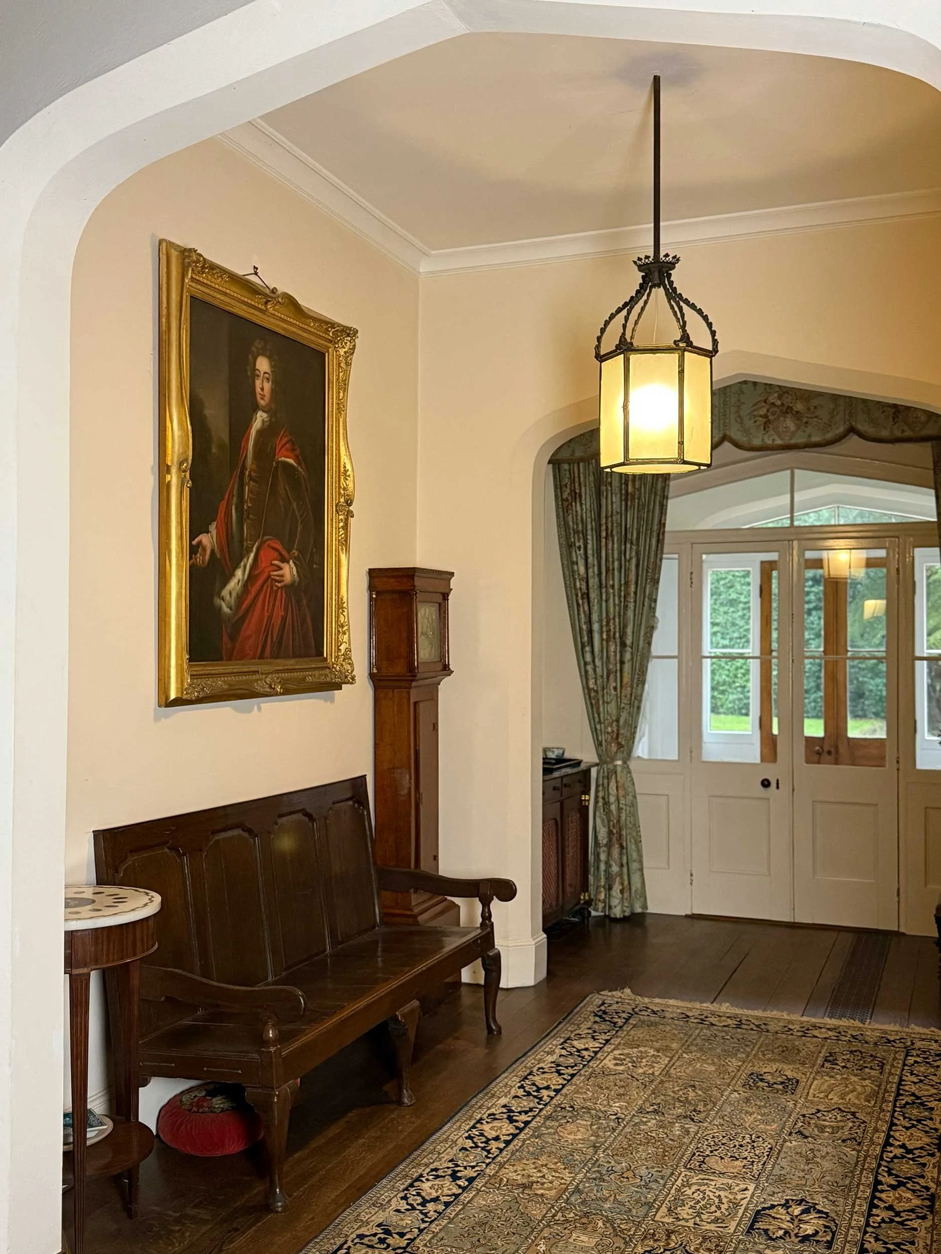 Interior of a room with a wooden bench, a round side table, a large framed portrait painting on the wall, a vintage-style hanging light fixture, and a door with windows leading outside, with curtains and a patterned rug on the floor.