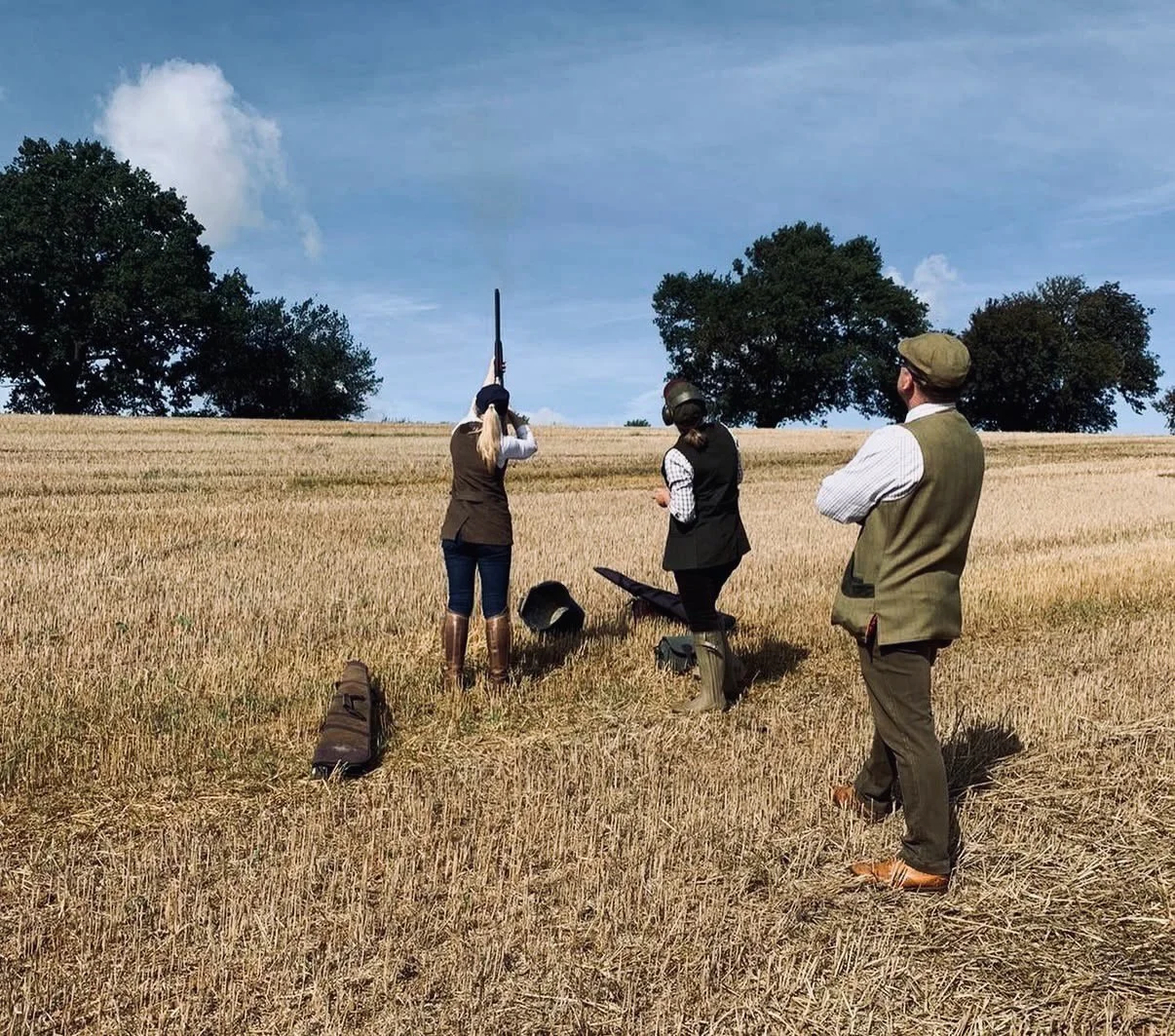 Group of three people in vintage clothing, standing in a wheat field, engaging in a historical reenactment with one person shooting a rifle. The others are watching; two are wearing caps and thematic attire. The scene appears to be in a rural area with trees under a partly cloudy sky.