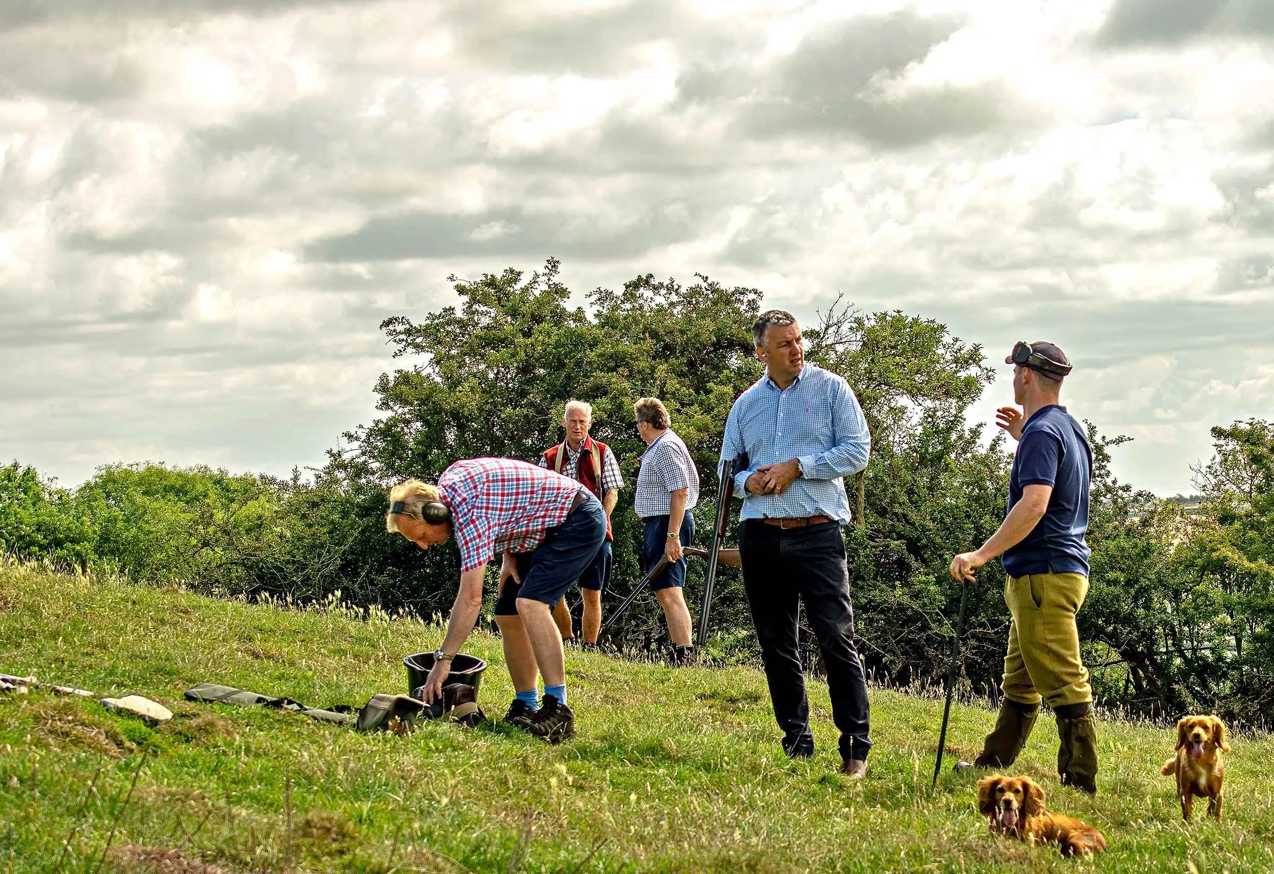 Group of five men outdoors in a grassy field with bushes and trees, some with dog companions. One man bends down, another stands with a cane, and they are engaged in conversation, with a cloudy sky overhead.