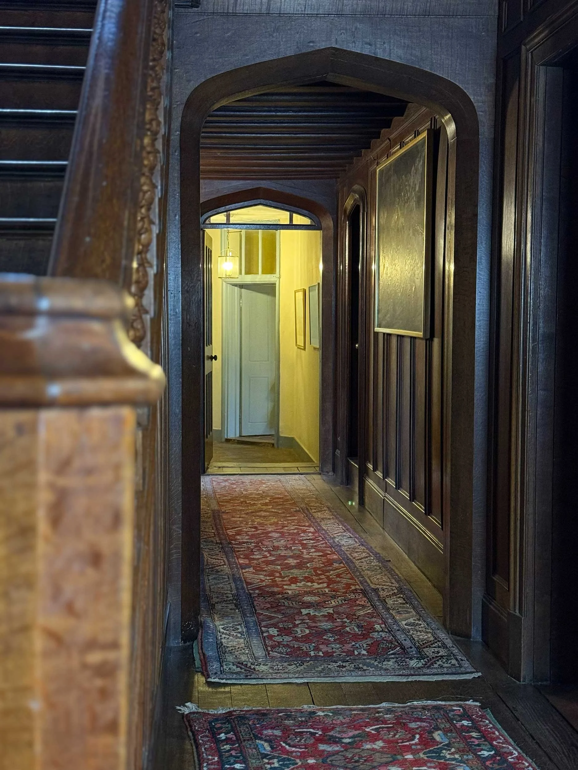 A hallway with wooden paneling on the walls, a patterned runner rug, and a view towards a yellow-lit room with a hanging light fixture and an open door.