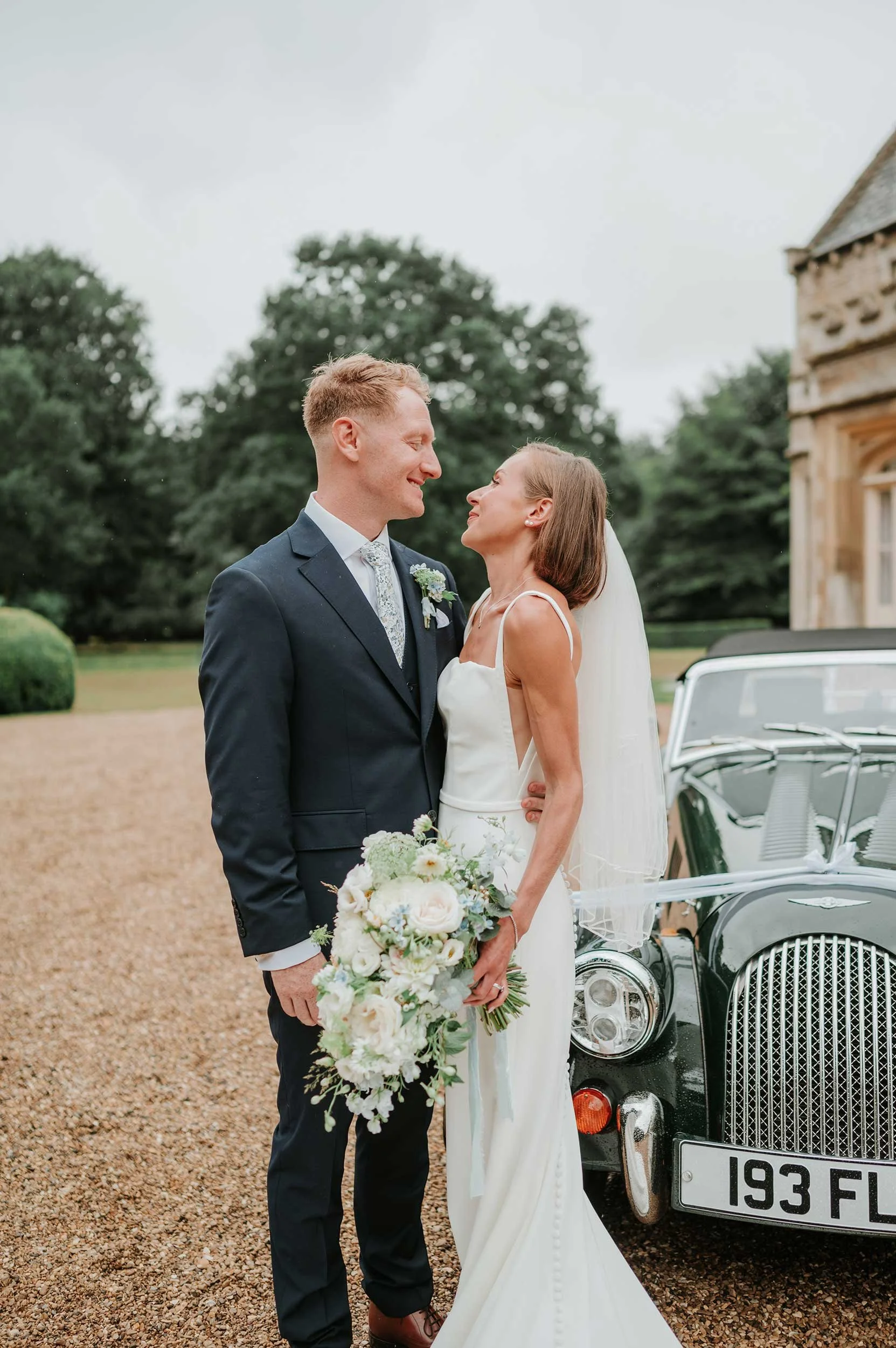 A bride and groom stand close together outdoors, looking into each other's eyes, near a vintage black car with a wedding ribbon, on a gravel driveway with trees and a stone building in the background.