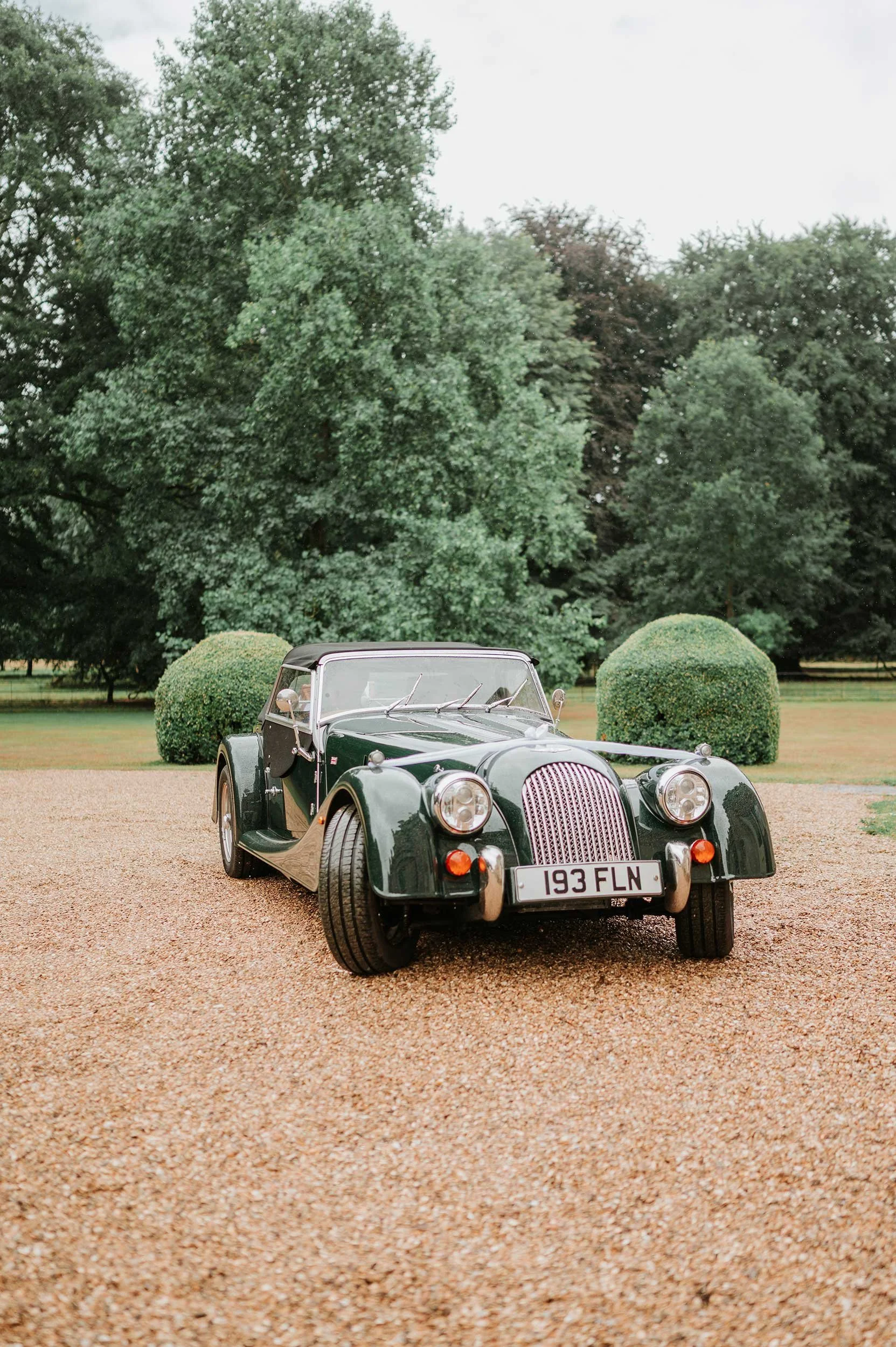 A vintage black convertible car parked on a gravel driveway with green trees and bushes in the background.