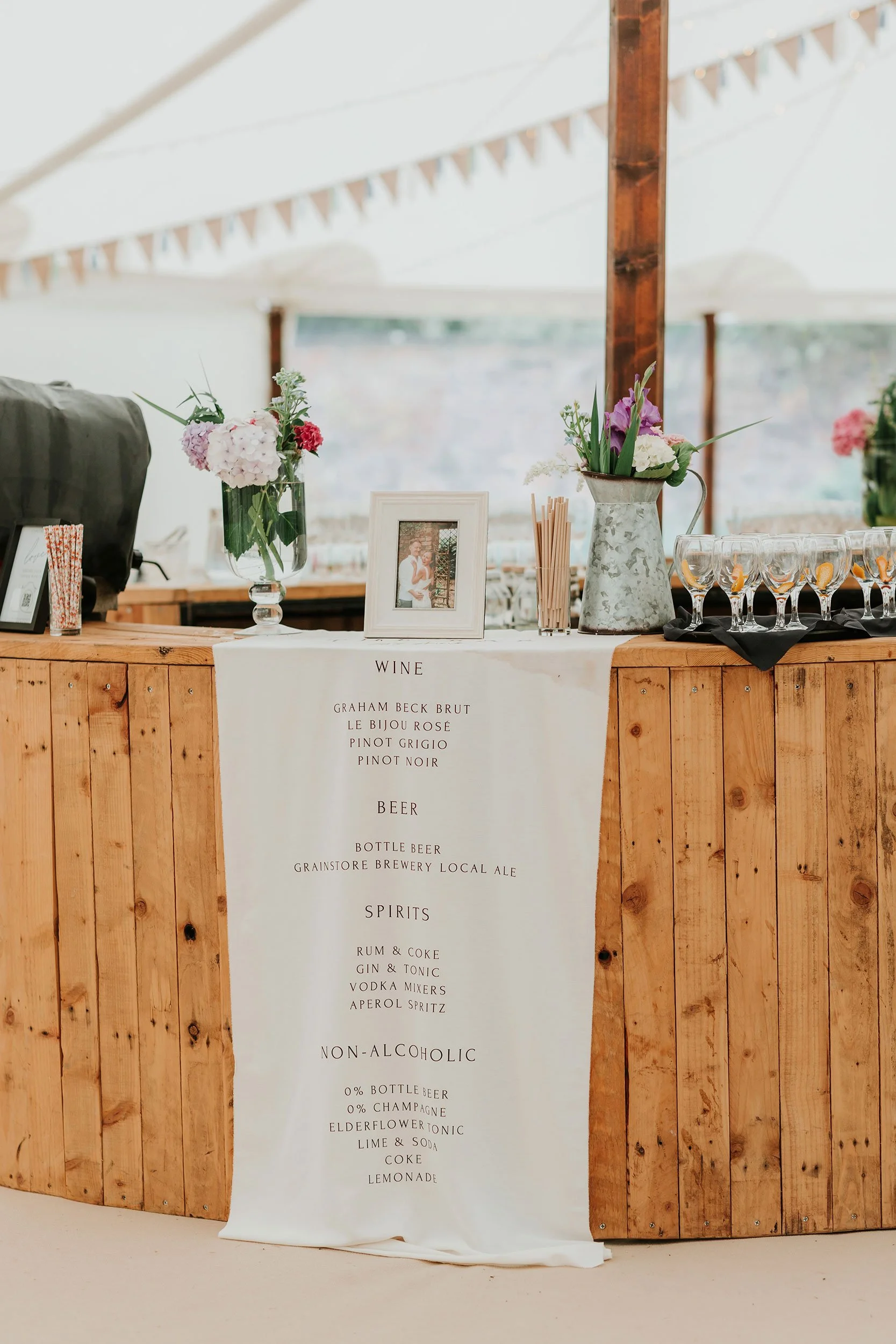 Wedding bar menu on white cloth with flowers and framed photograph on wooden counter.