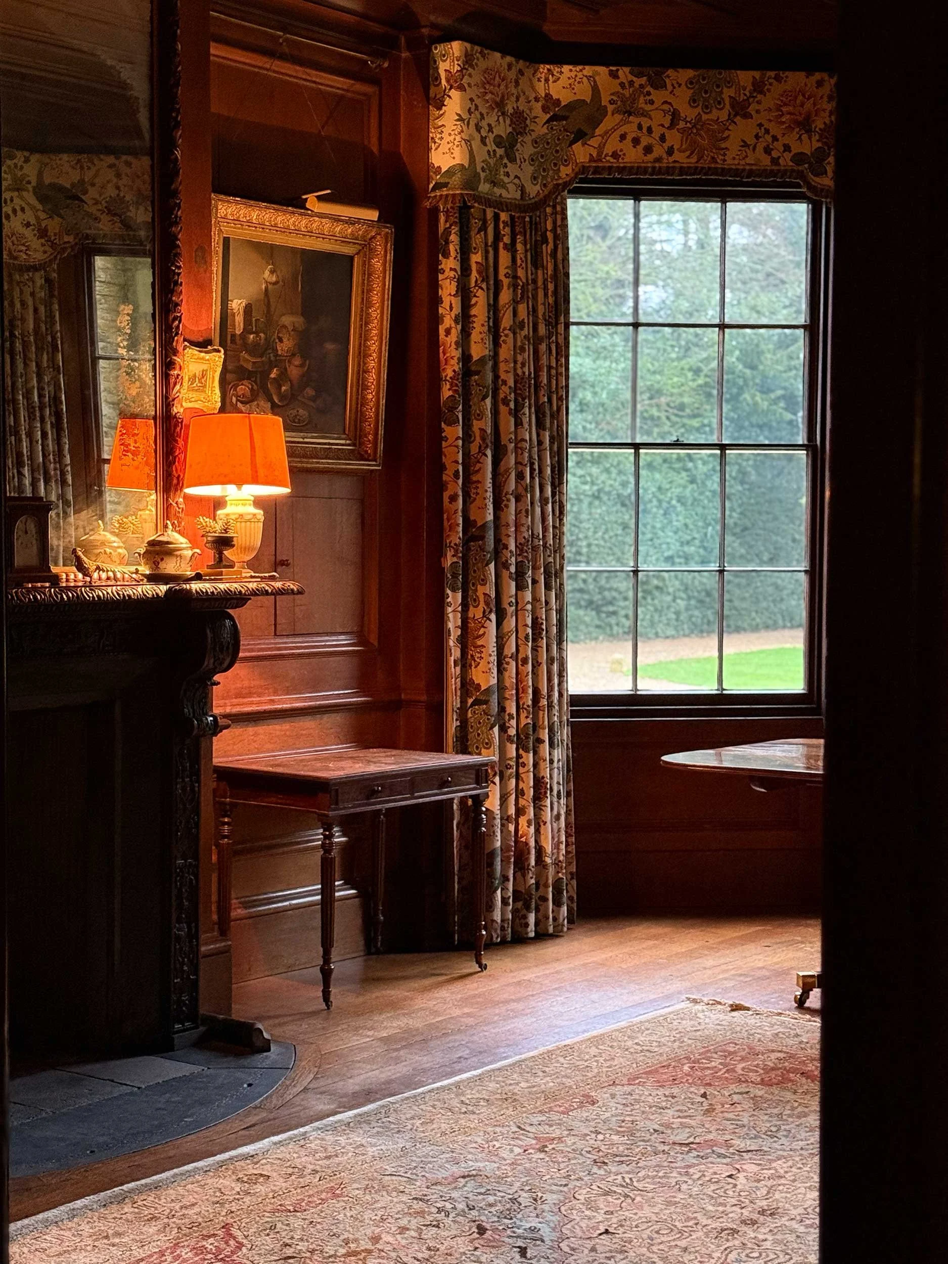 A cozy interior room featuring a large window with patterned curtains, a wooden table, a framed painting on the wall, and a lamp casting warm light.