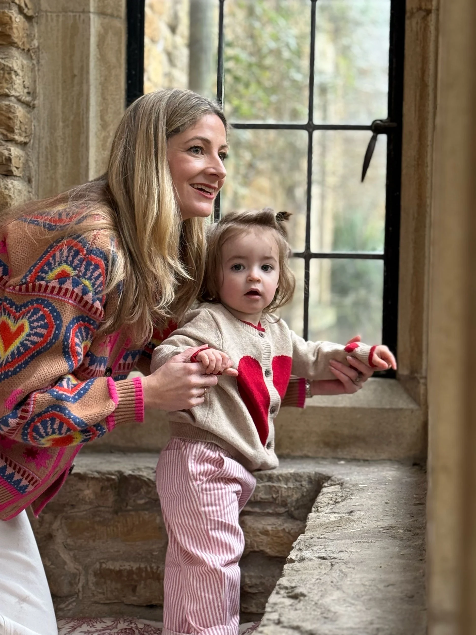 A woman and a young girl looking out of a window in . The woman is smiling, and the girl looks curious. The woman has long blonde hair and is wearing a colorful patterned sweater. The girl has brown hair tied with small pigtails and is wearing a beige sweater with a red heart and pink striped pants.
