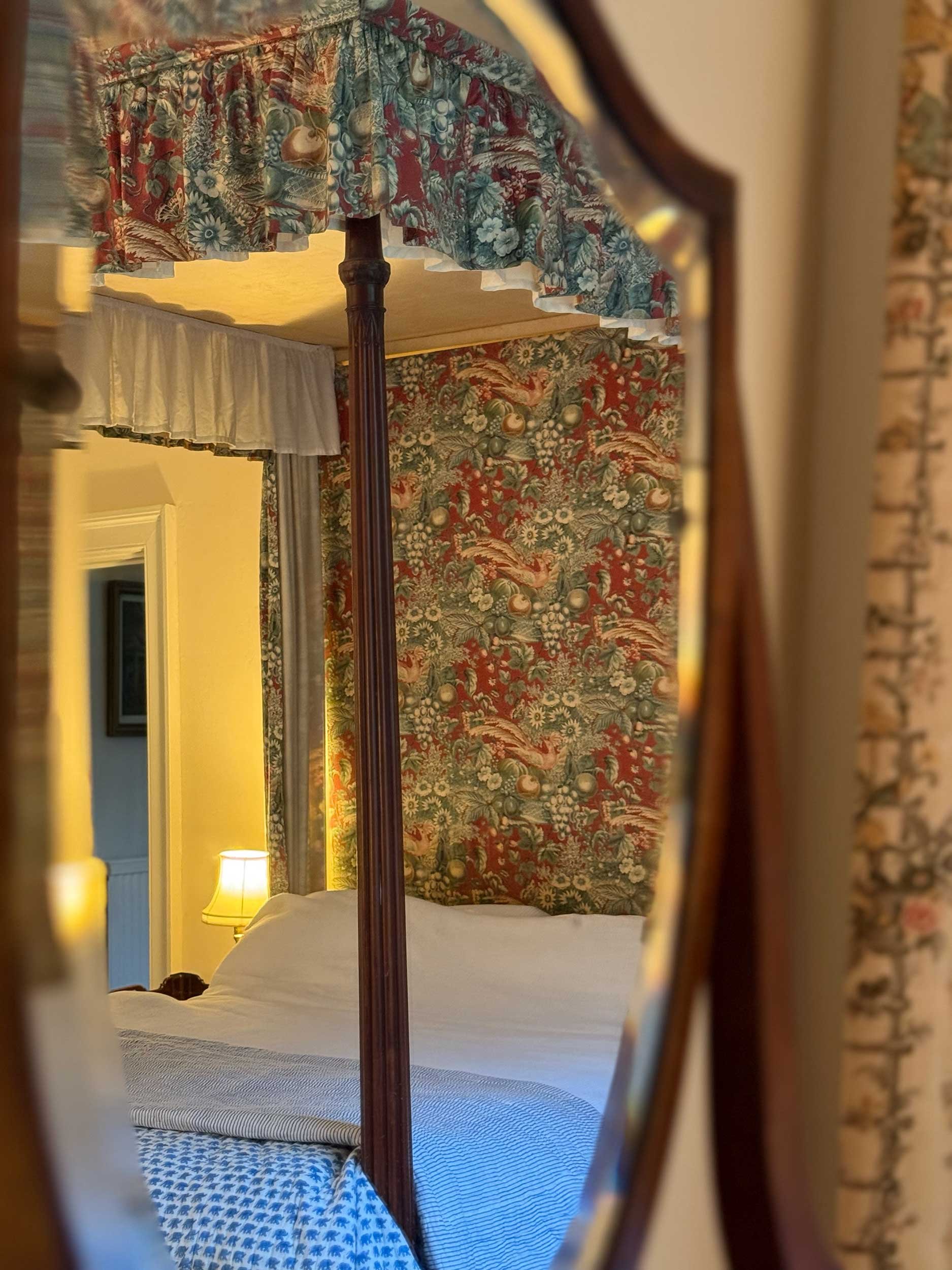 Reflection of a bedroom with a canopy bed, patterned wallpaper, a lamp, and curtains, seen through a decorative mirror.