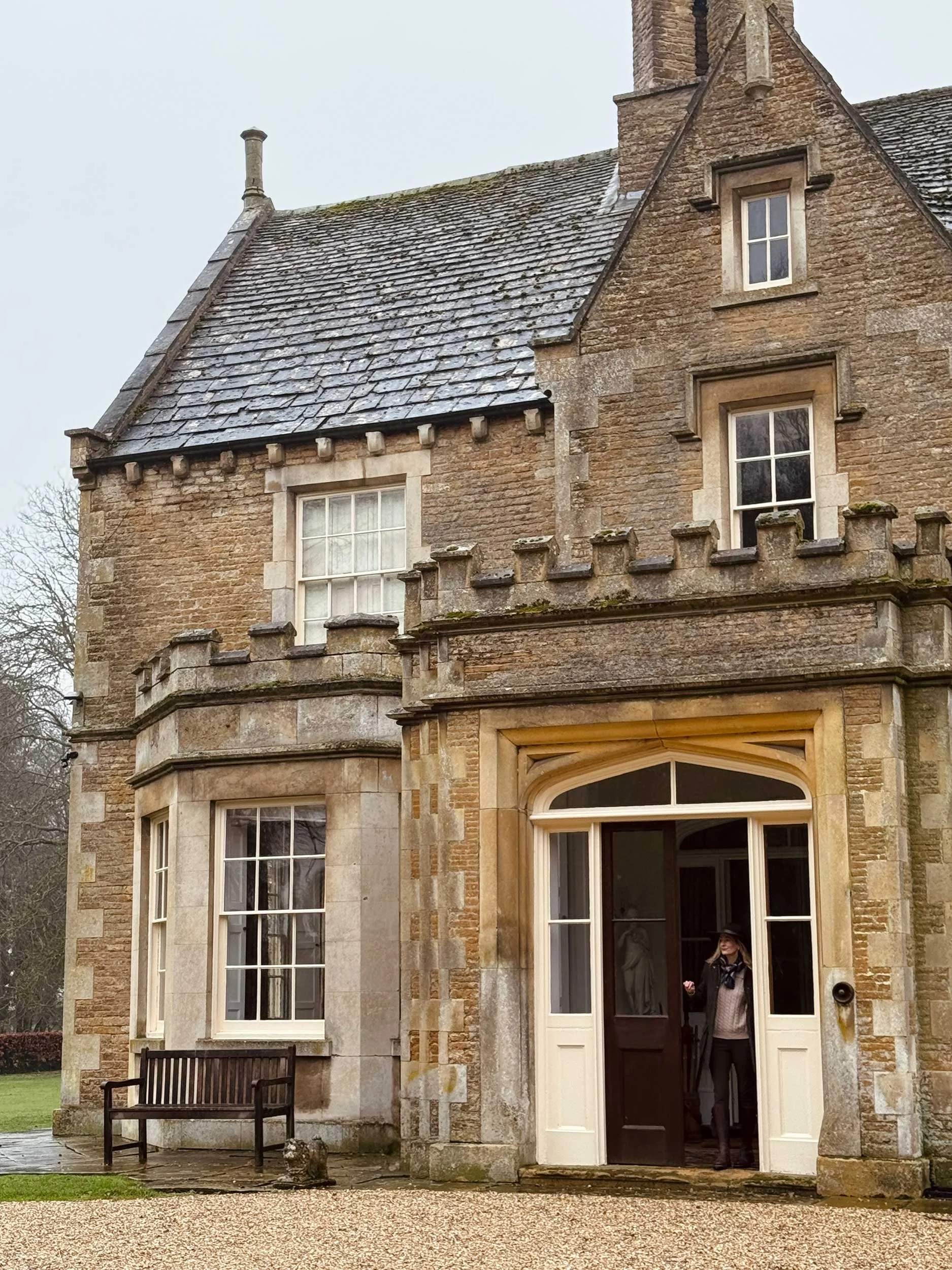 A historic stone house with a woman standing in the doorway, a bench outside, and a gravel path in front.