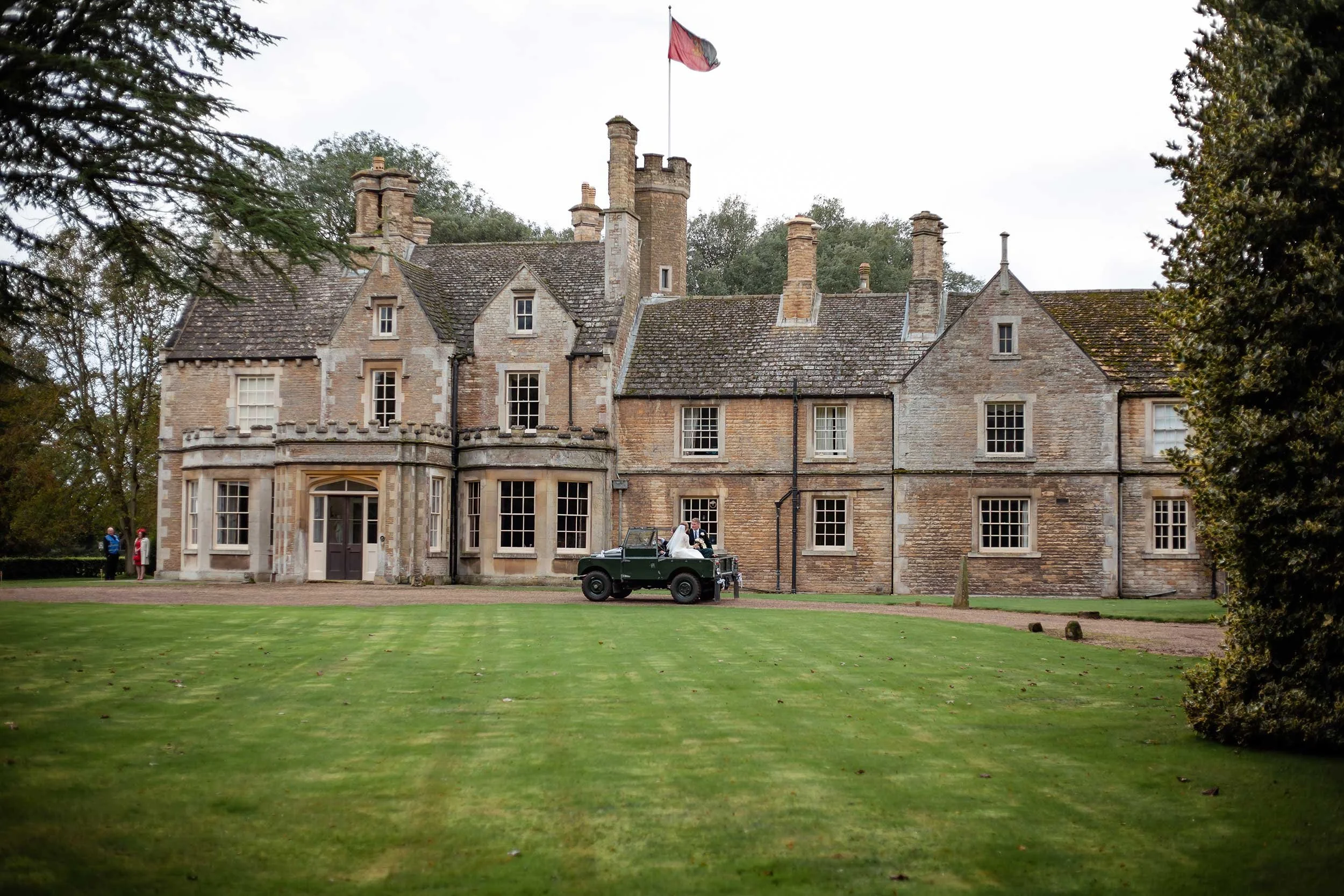 A wedding couple in wedding attire sitting on a vintage green car parked in front of a historic stone mansion with multiple chimneys and a flag on top, set on a well-maintained lawn with trees in the background.