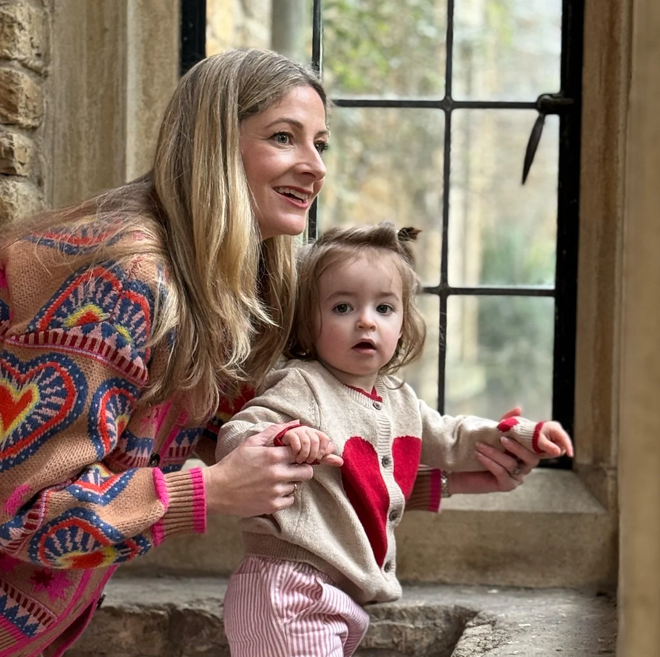A woman with long blonde hair and a young girl with curly hair are looking out of a window. The woman is holding the girl's hands and smiling, while the girl has a curious expression. The woman is wearing a colorful patterned sweater, and the girl is wearing a beige jacket with a red heart on it.