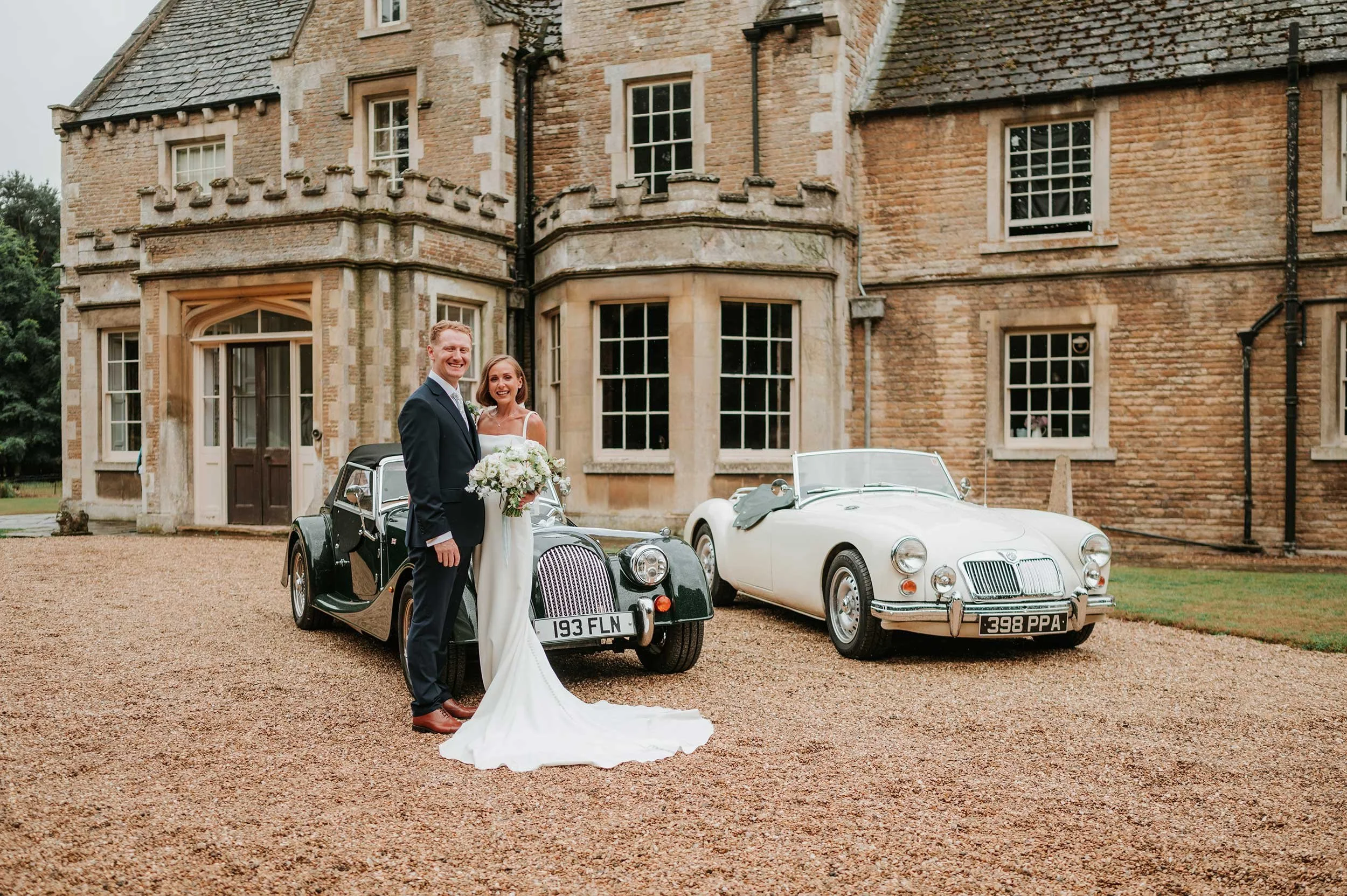 A newlywed couple standing on a gravel driveway in front of an old stone mansion, with two vintage cars parked behind them, one black and one white, celebrating their wedding day.