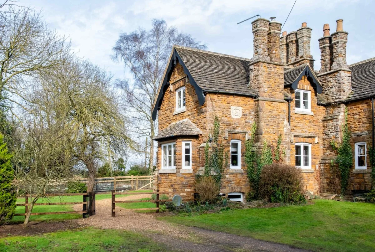 A stone cottage with multiple tall chimneys, small dormer windows, and ivy climbing its exterior, surrounded by a garden and trees.