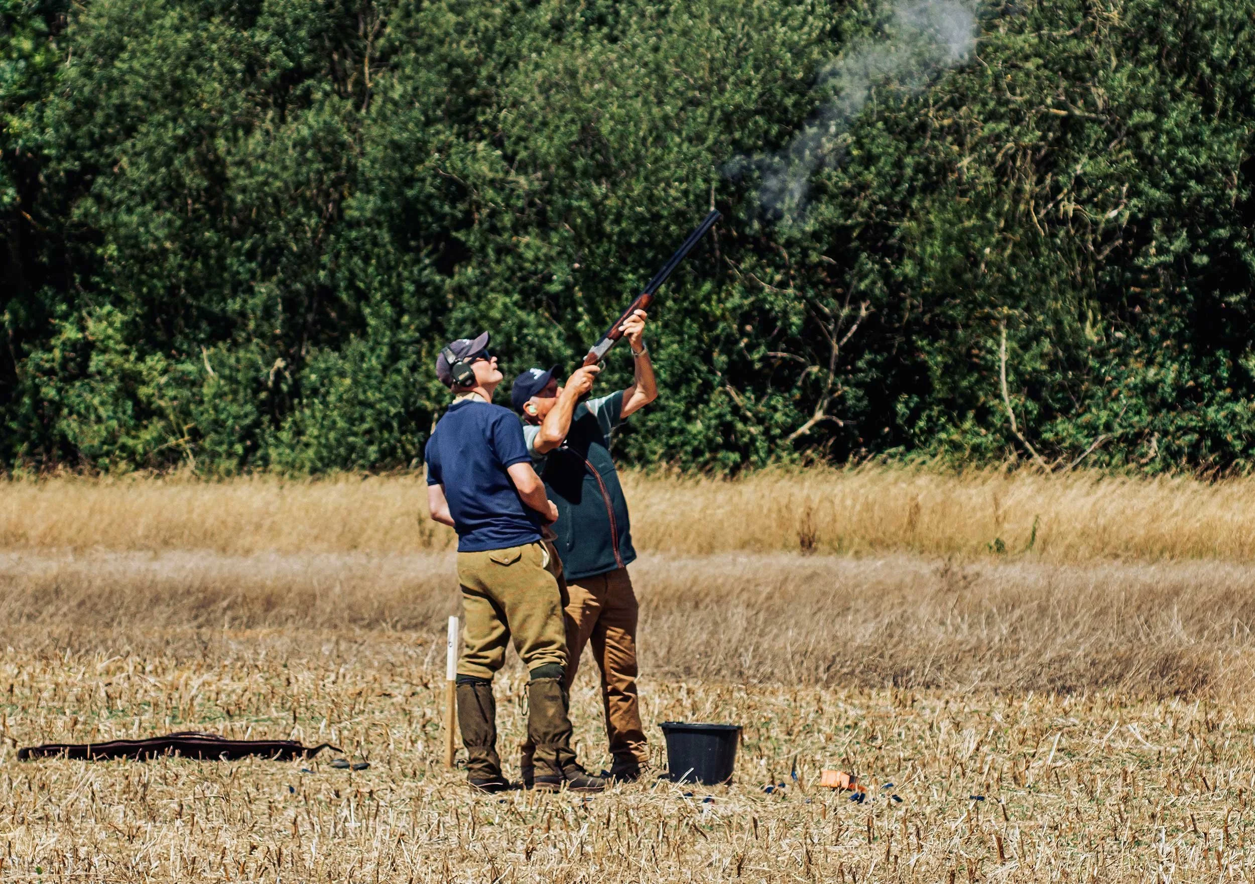 Two men stand in a field, one aiming a shotgun upwards while the other watches. Smoke is coming out of the shotgun. There are trees in the background.