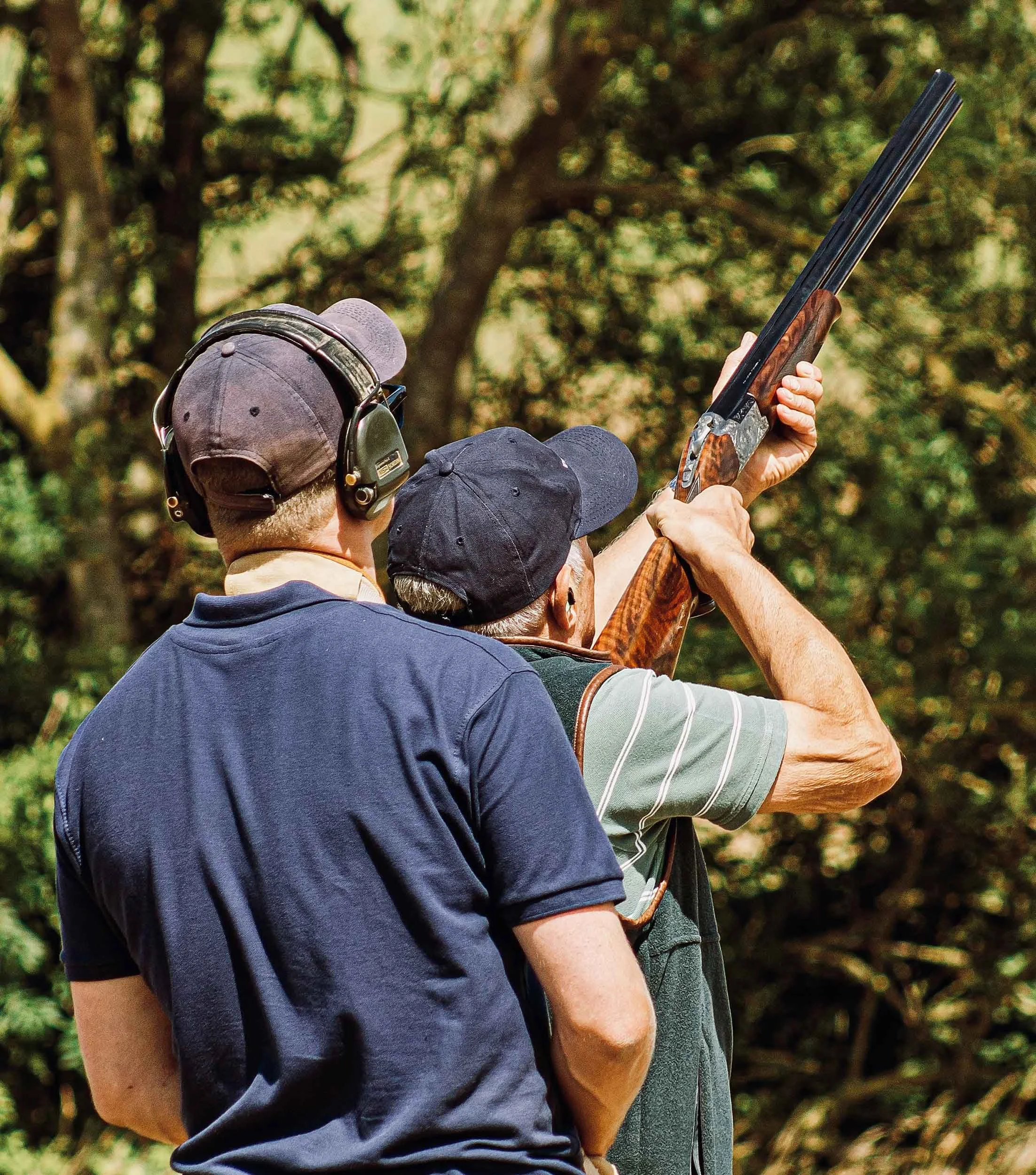Two men are at a shooting range, one aiming a shotgun with the other assisting. The man shooting is wearing a dark cap and resting the shotgun on his shoulder, while the other man is wearing headphones and a baseball cap, standing close by.
