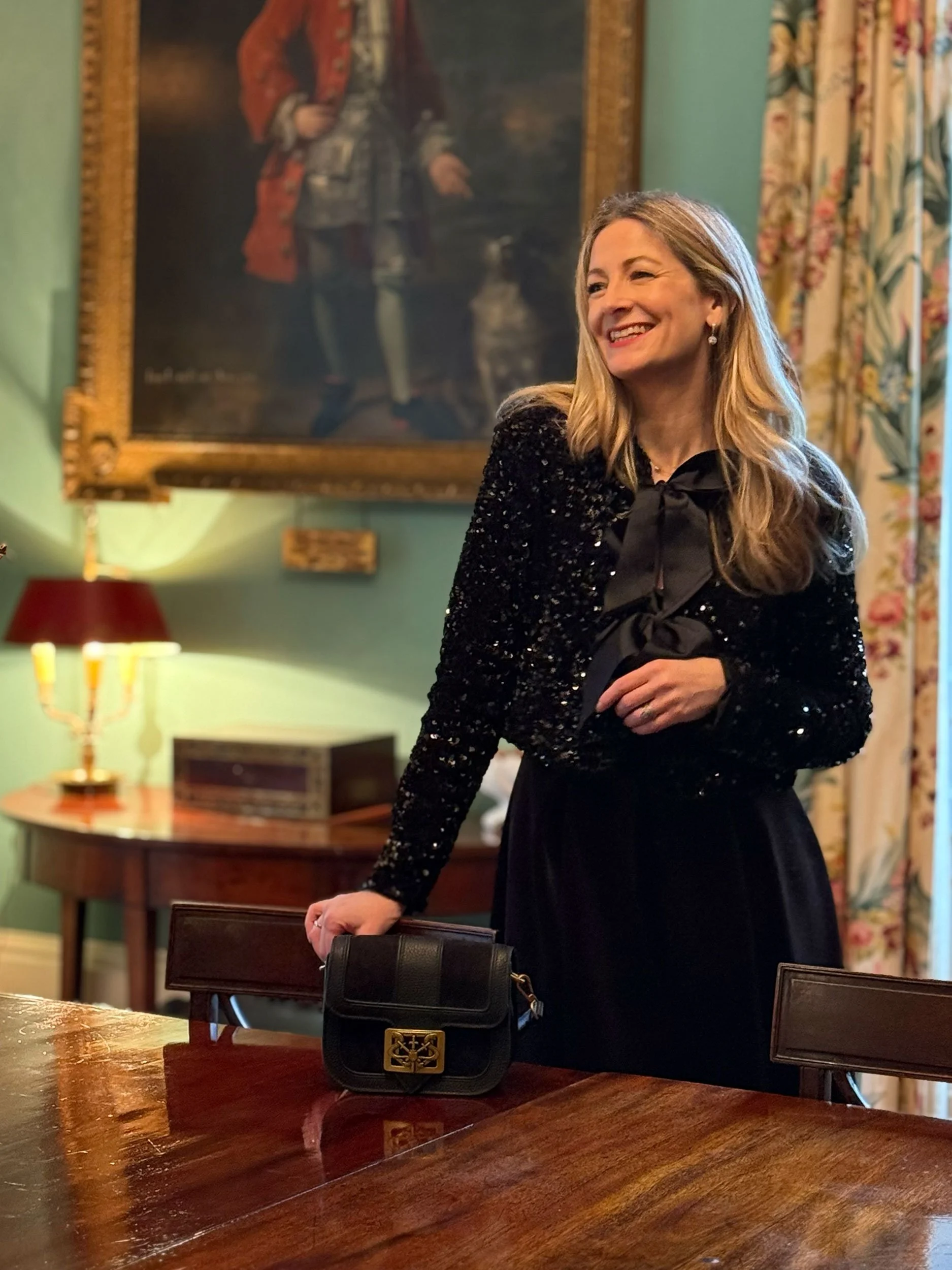 A smiling woman in a black outfit standing next to a wooden table with a black purse on it, in a room with green walls, a large portrait painting, floral curtains, and a lamp in the background.