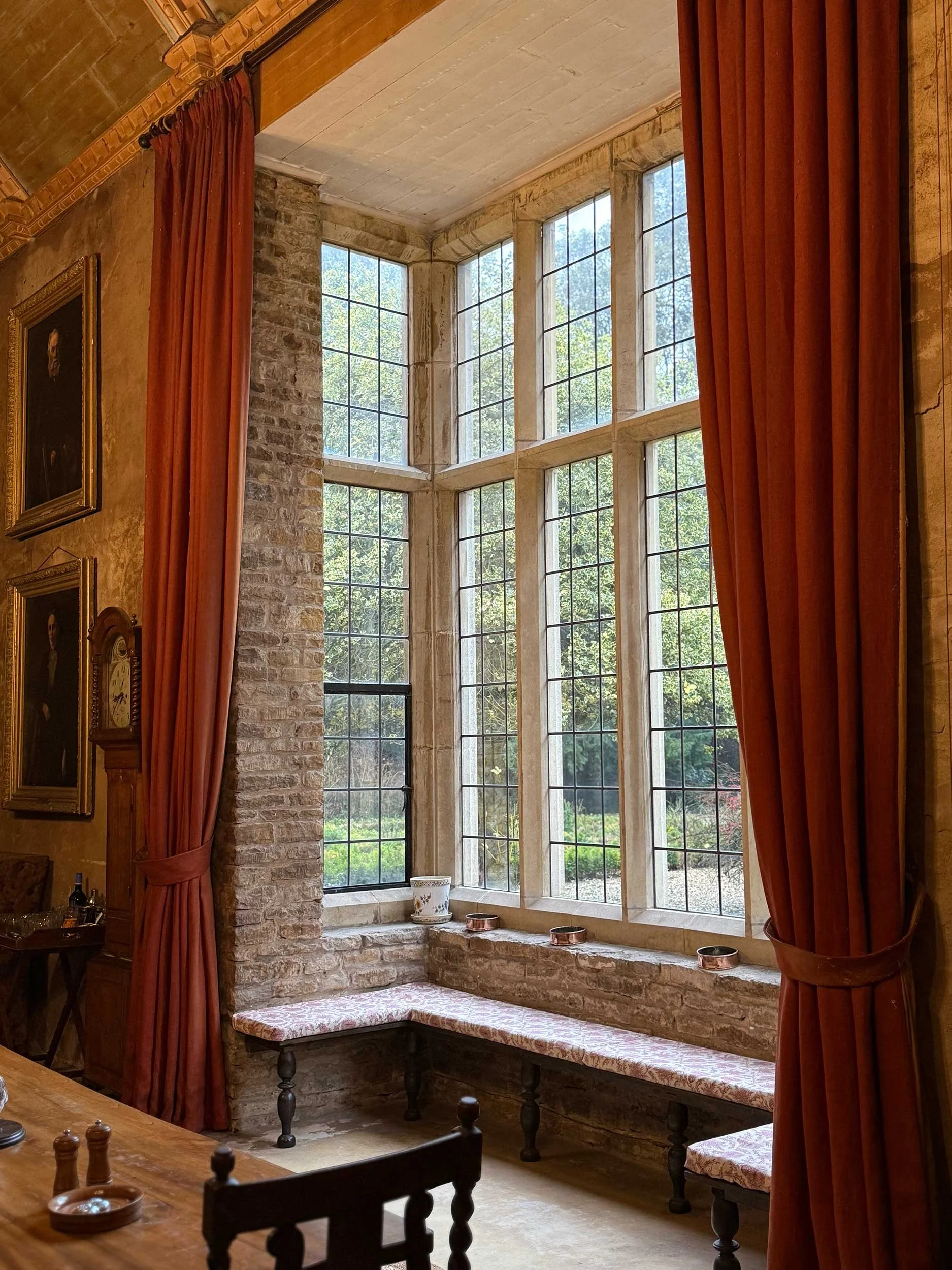 Sunlit corner of a room with large multi-pane windows, brick wall, wooden shelf, and a cushioned window seat with patterned fabric, red curtains, and decorative items.