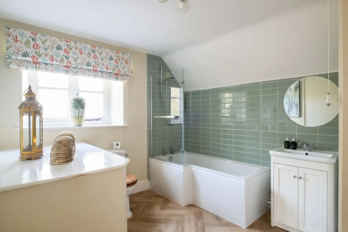 Bathroom with a window, a bathtub with a glass shower screen, a white vanity with a round mirror, and decorative items including a lantern and a vase with plants.