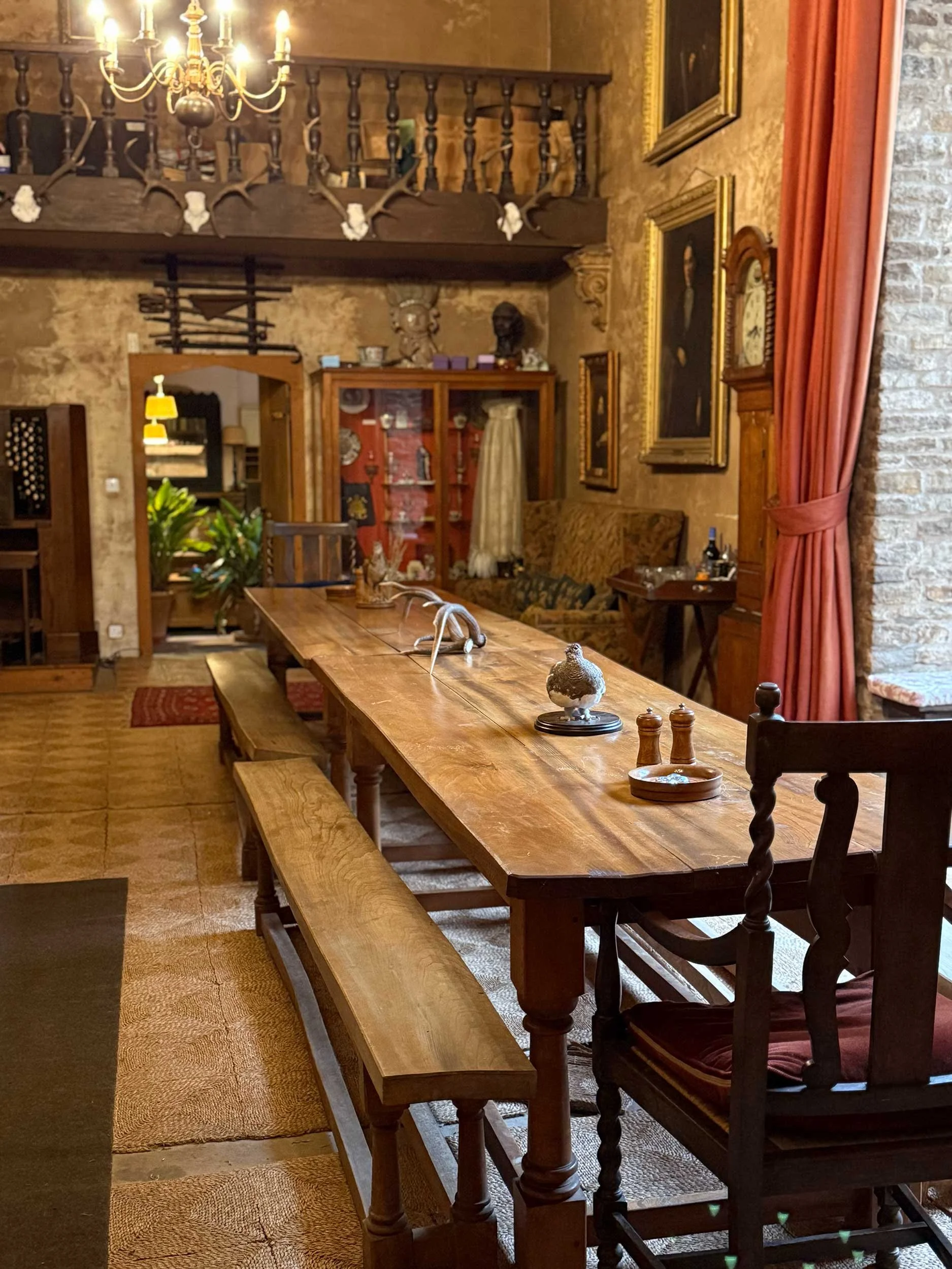 A cozy, rustic dining room with a long wooden table and benches, vintage furniture, artwork on brick walls, and a chandelier hanging from the ceiling.