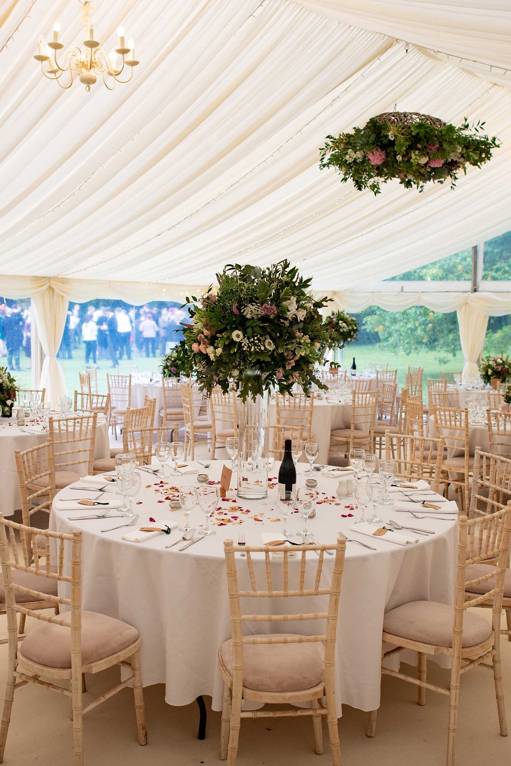 Wedding reception setup inside a canopy tent with a round table decorated with a large floral centerpiece, surrounded by beige chairs, wine glasses, and scattered rose petals.