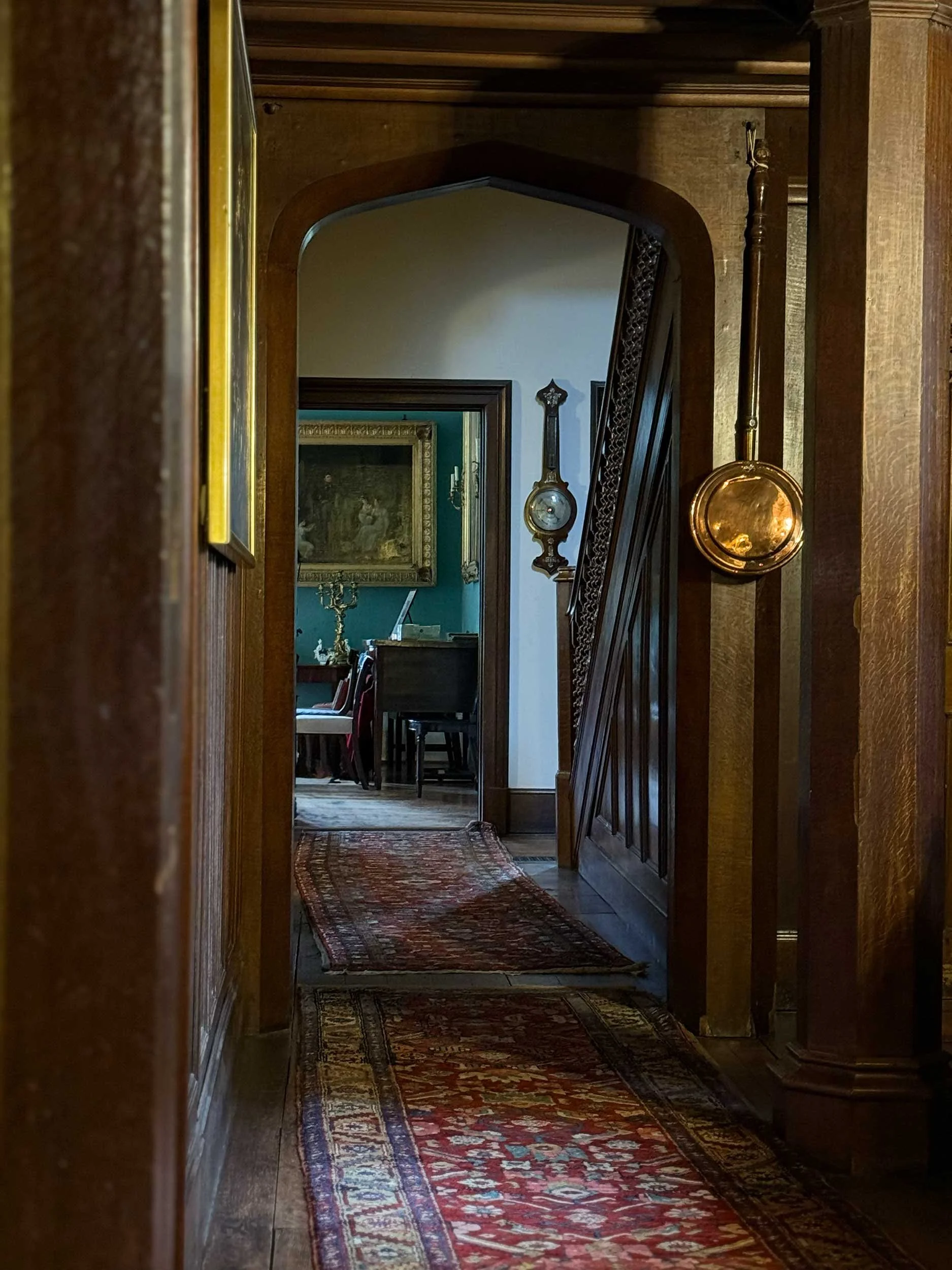 View of a hallway with antique wooden paneling and Persian runners, leading to a dining room with green walls, a framed painting, and a chandelier.