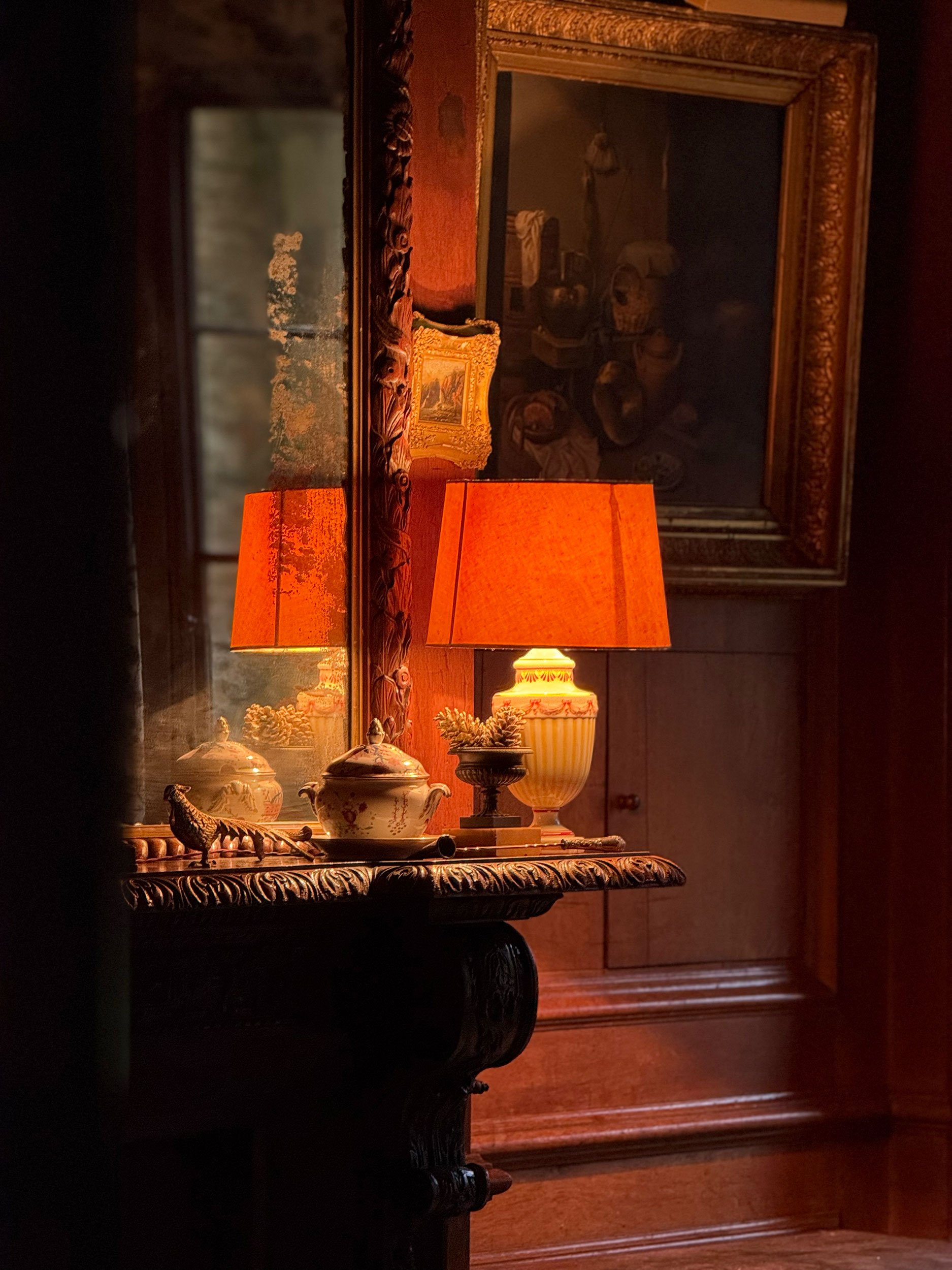 A cozy corner of a room with a wooden dresser, a large mirror, two orange lamps, and decorative teacups and figurines on the dresser. A framed still life painting hangs on the wall.