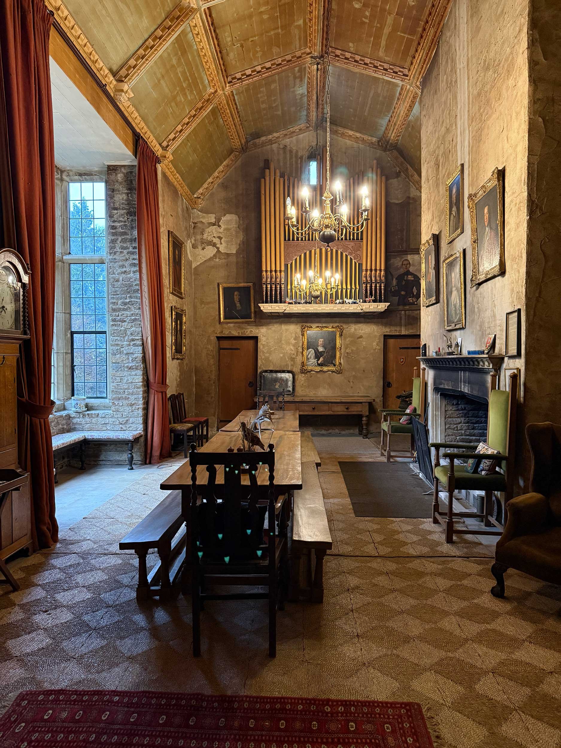 Historic room with high, gold-accented vaulted ceiling and stone walls. Large window with red curtains, antique chairs, a fireplace with framed portraits above, a long wooden table, and a pipe organ at the far end illuminated by chandeliers.