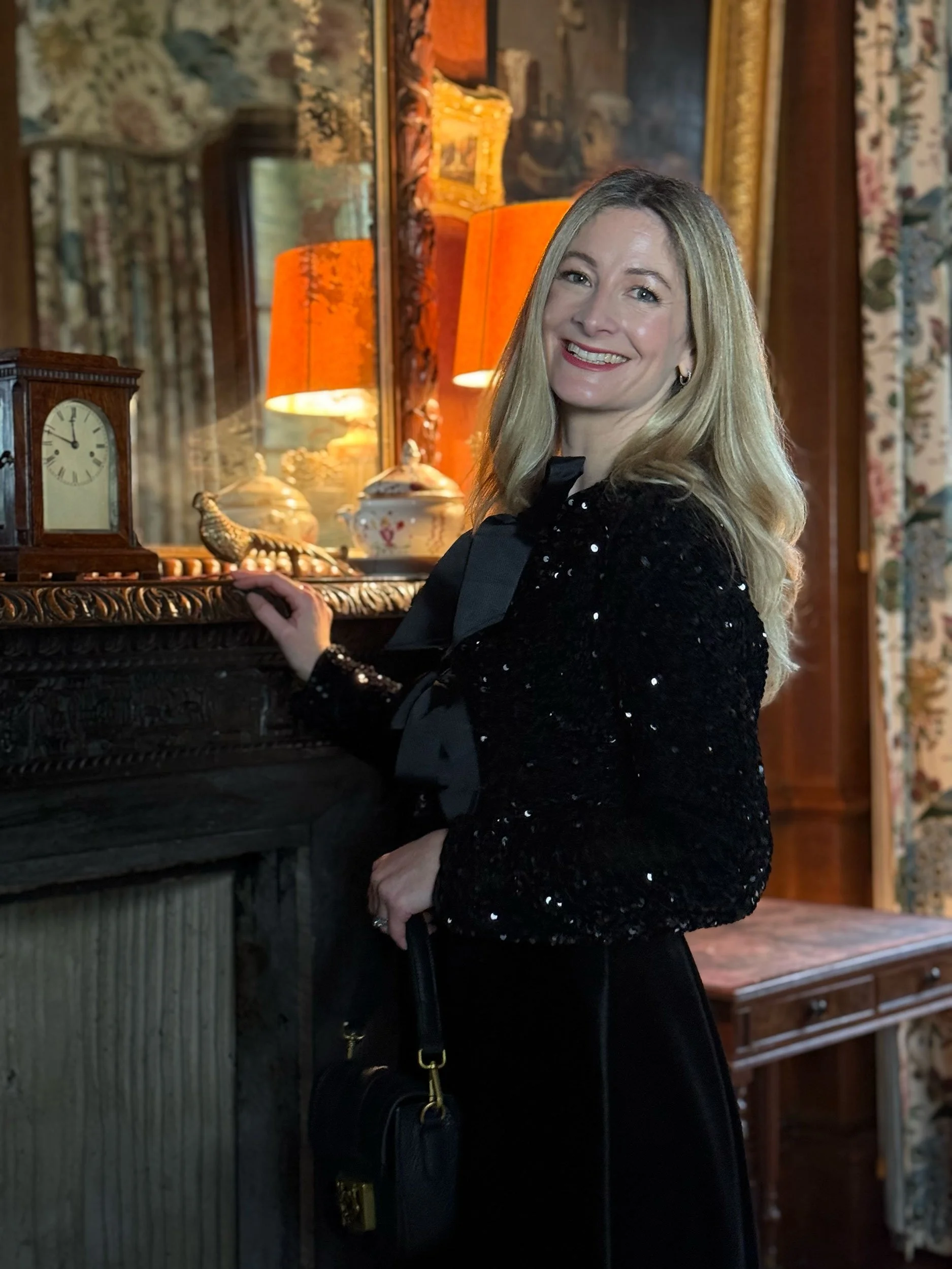 A woman with blonde hair, smiling, dressed in black, standing in a room with vintage decor, next to a mantle with a clock, lamps, and decorative items.