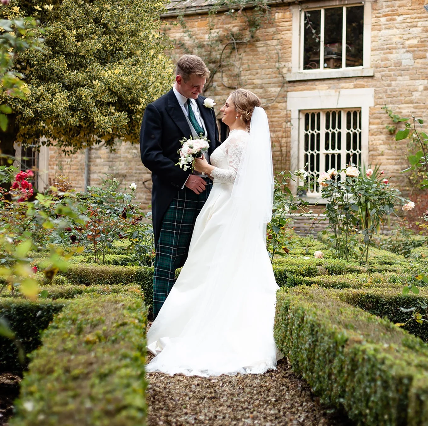 A couple dressed in wedding attire stands in a garden, gazing at each other, with the woman holding a bouquet of flowers and a historic brick building in the background.