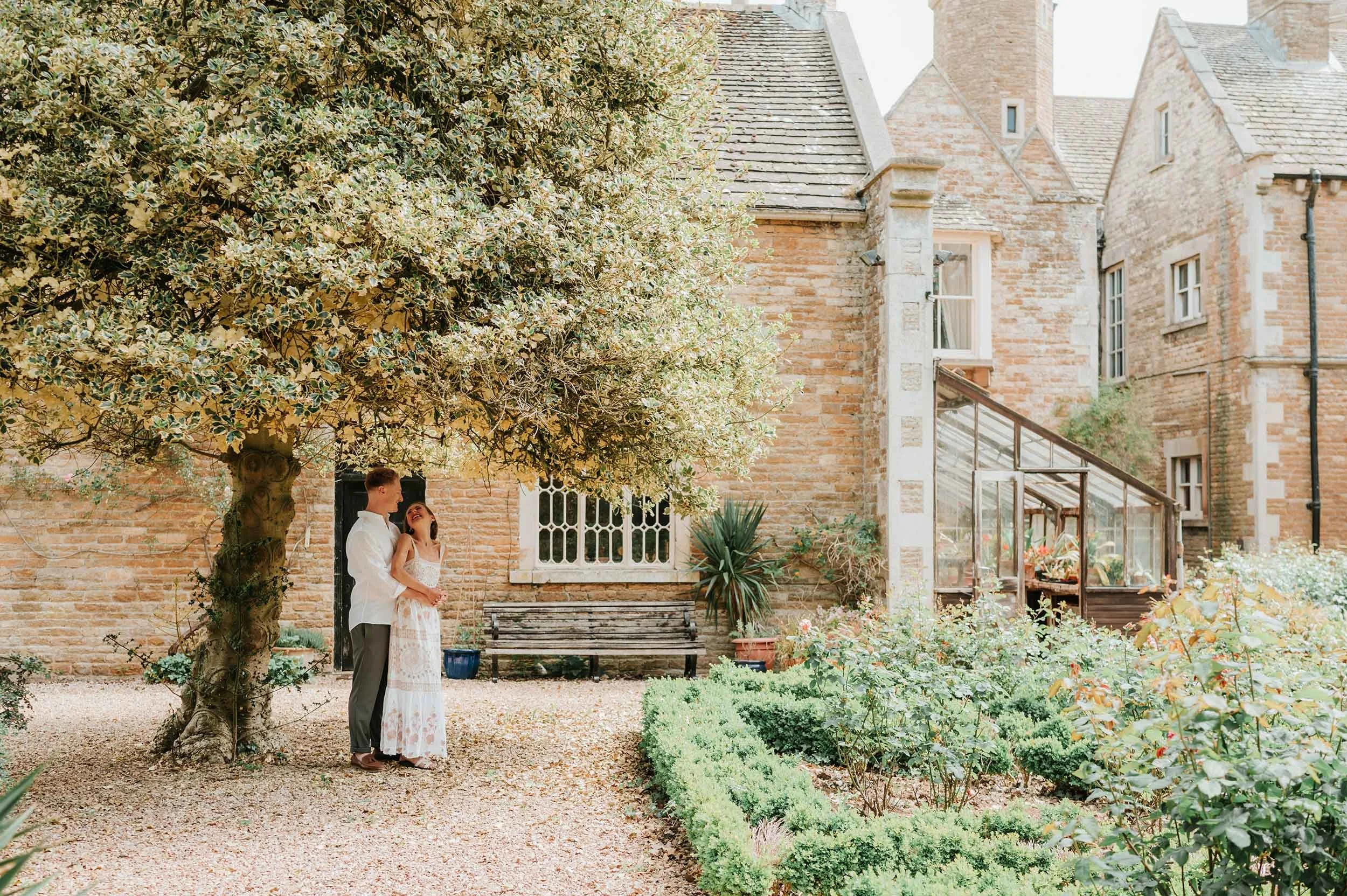 A couple standing under a large leafy tree in a garden courtyard next to an old brick building with a greenhouse attached.
