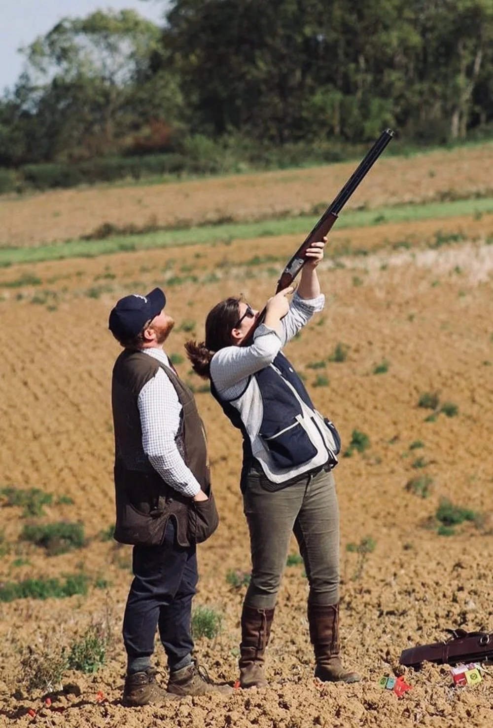 Two people standing in a field, with one person aiming a shotgun skyward, while the other observes. The field is brown and green, with trees in the background.