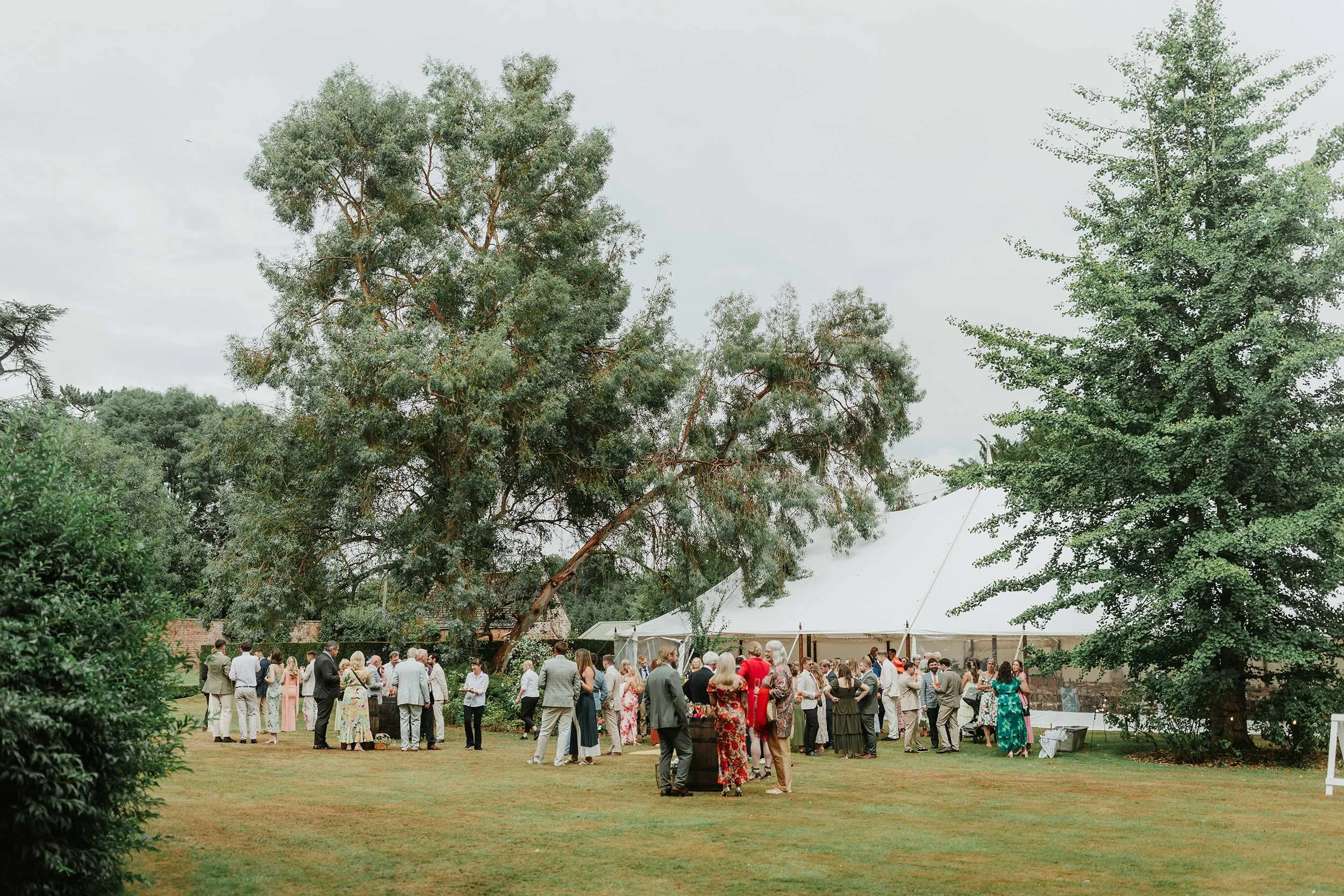 Outdoor wedding reception with guests mingling near a large white tent, surrounded by trees and a well-maintained grassy lawn.