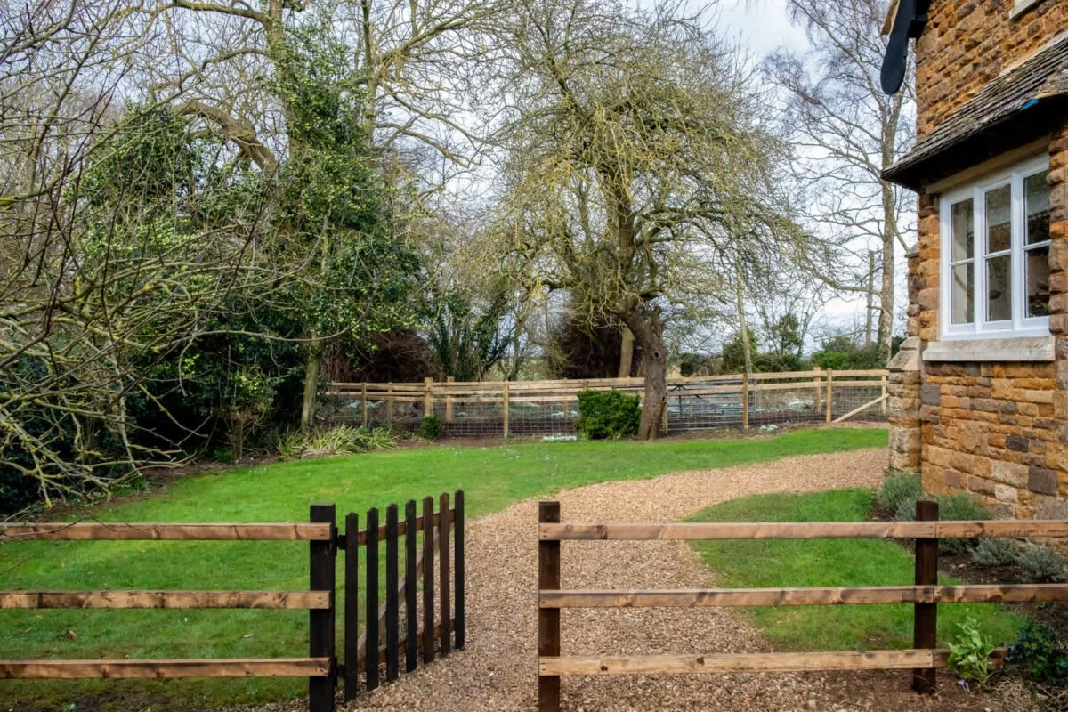 View of a fenced backyard with trees, a stone cottage on the right, a gravel path, and a lawn.