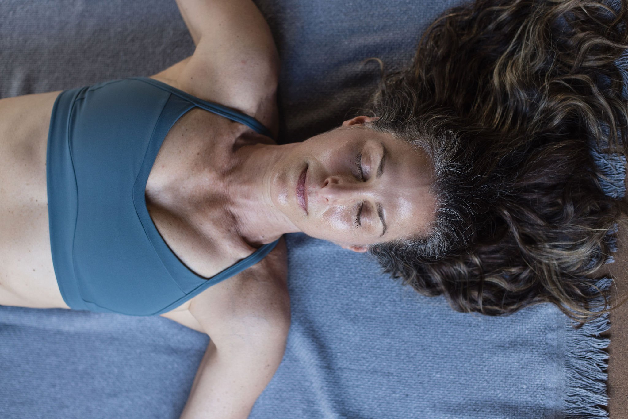 A woman with curly hair and freckles is lying on a gray yoga mat with her eyes closed, wearing a blue sports bra.