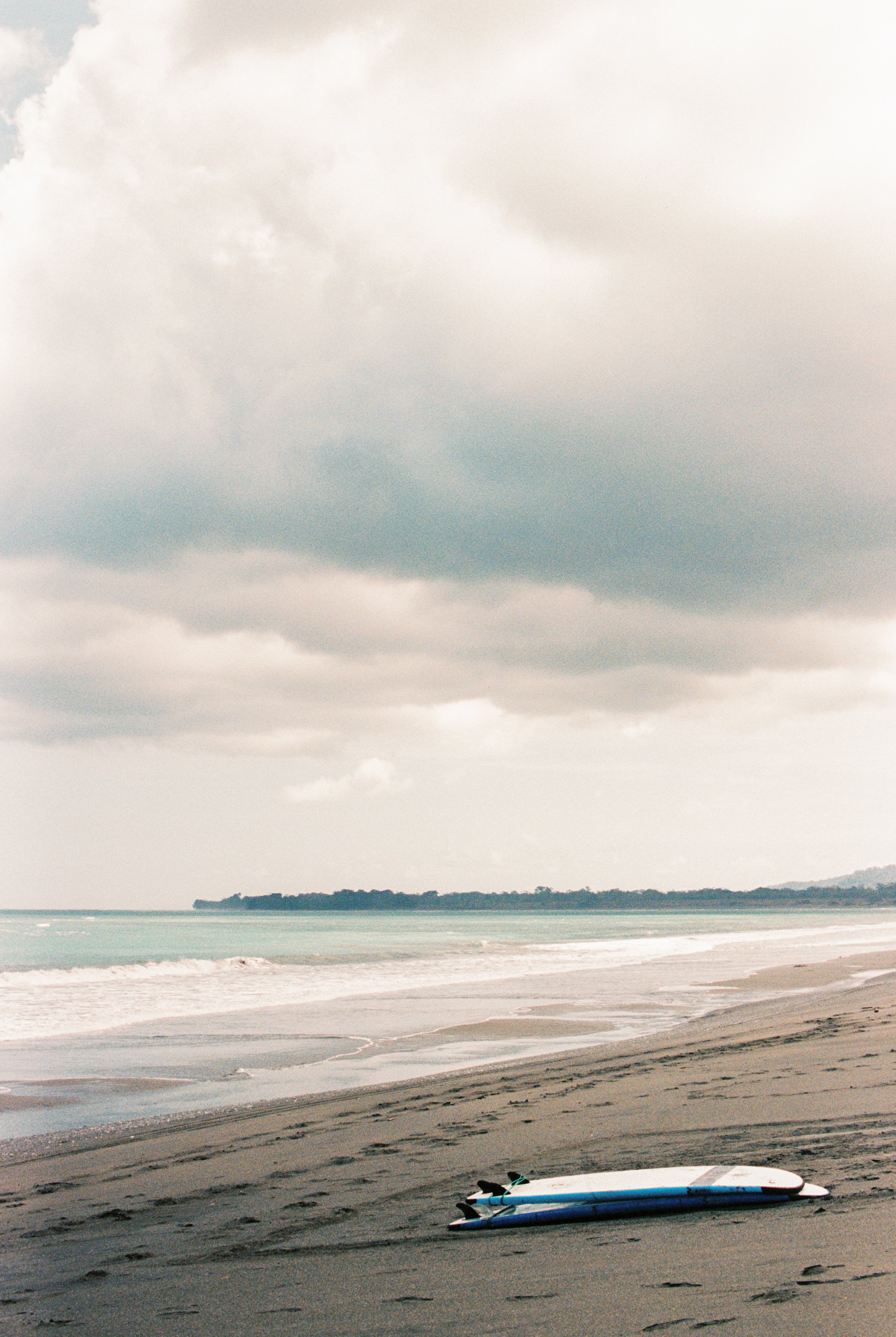 A beach with a surfboard and a few paddleboards lying on the sand near the water, with an overcast sky and a distant landmass across the ocean.