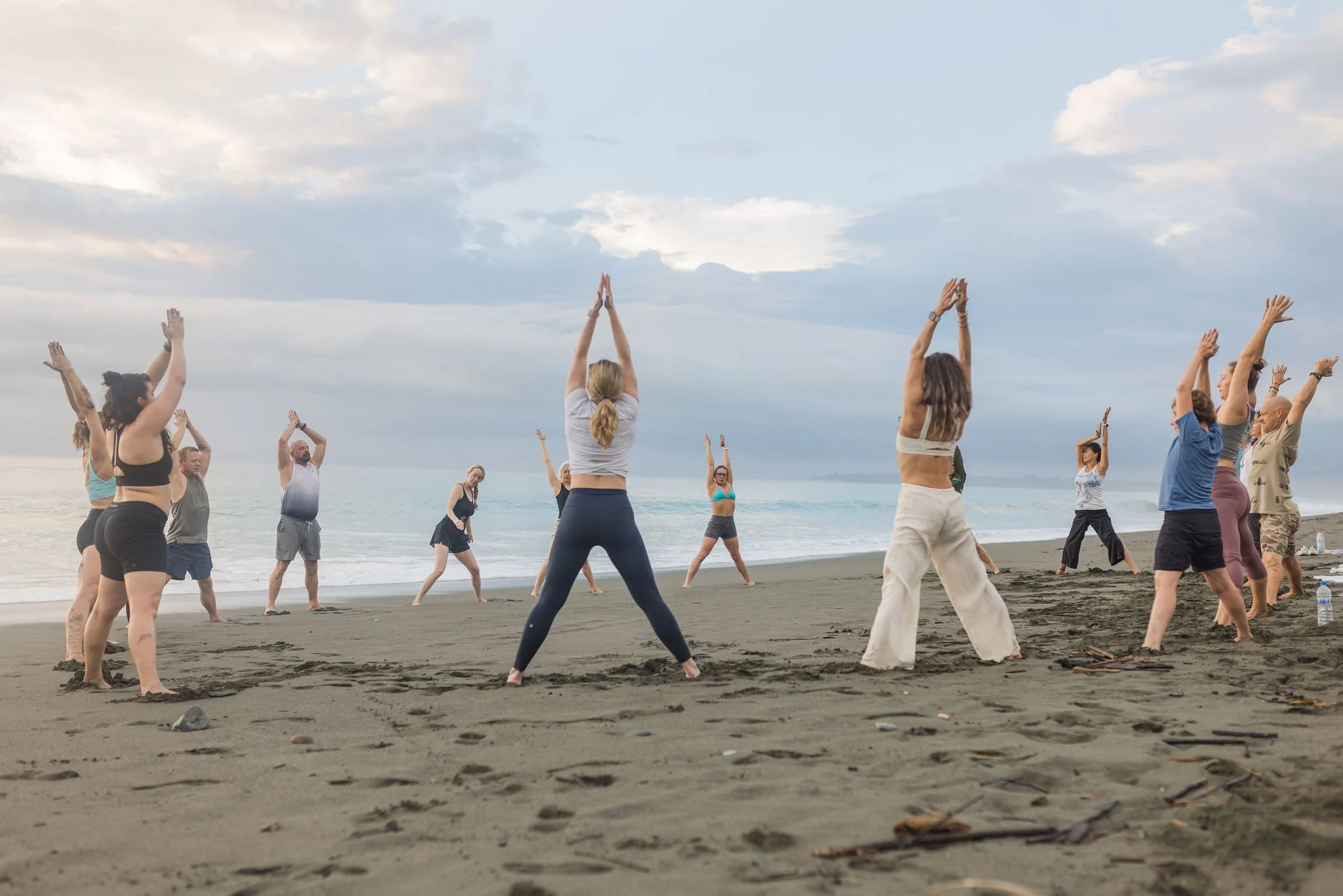 A group of people practicing yoga on a beach during sunset, standing in a circle with hands raised above their heads, facing the ocean.