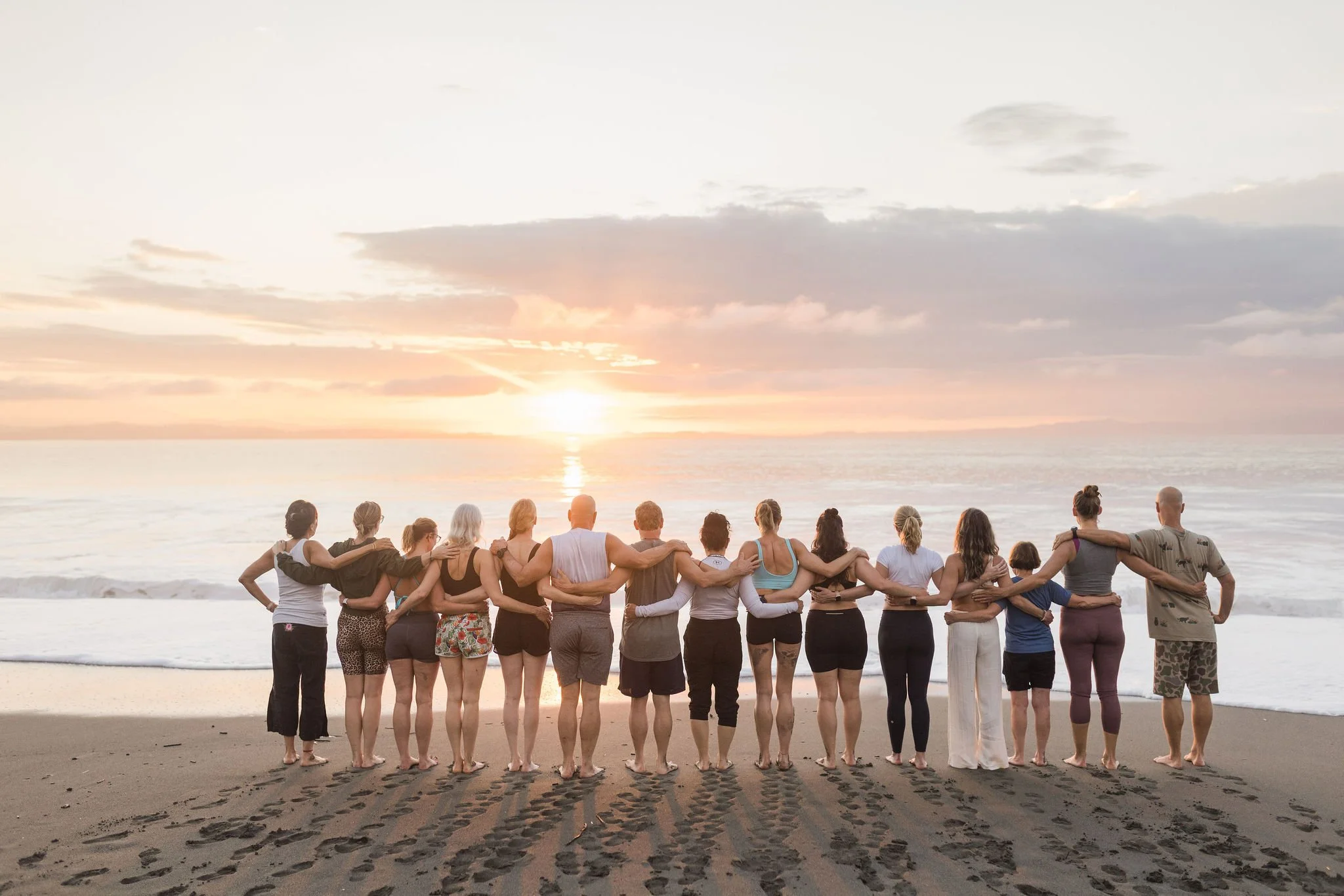 A diverse group of people standing in a line on the beach with their arms around each other, facing the ocean at sunset.