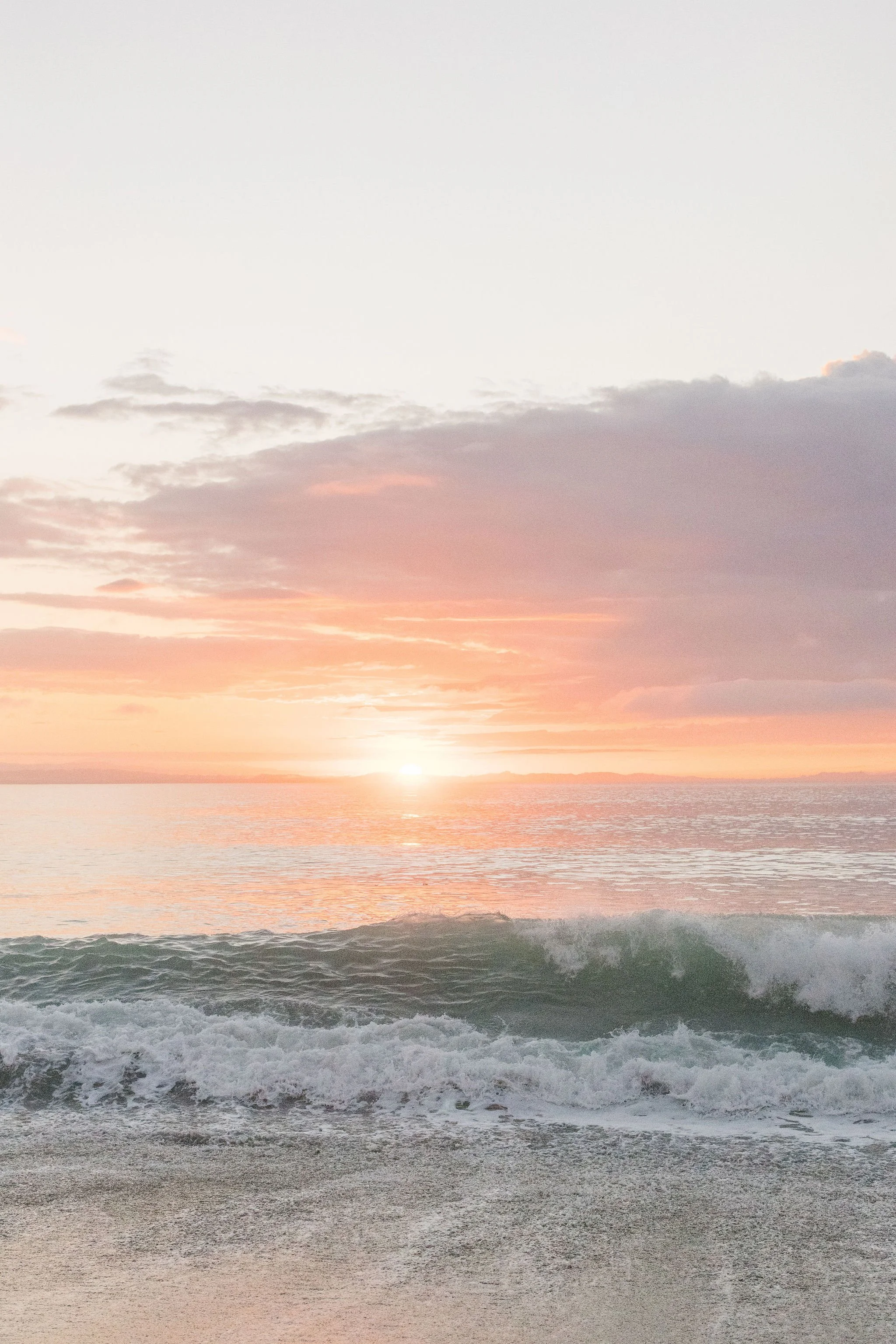 Sunset over the ocean with waves crashing on the shore, partly cloudy sky with pink and orange hues.