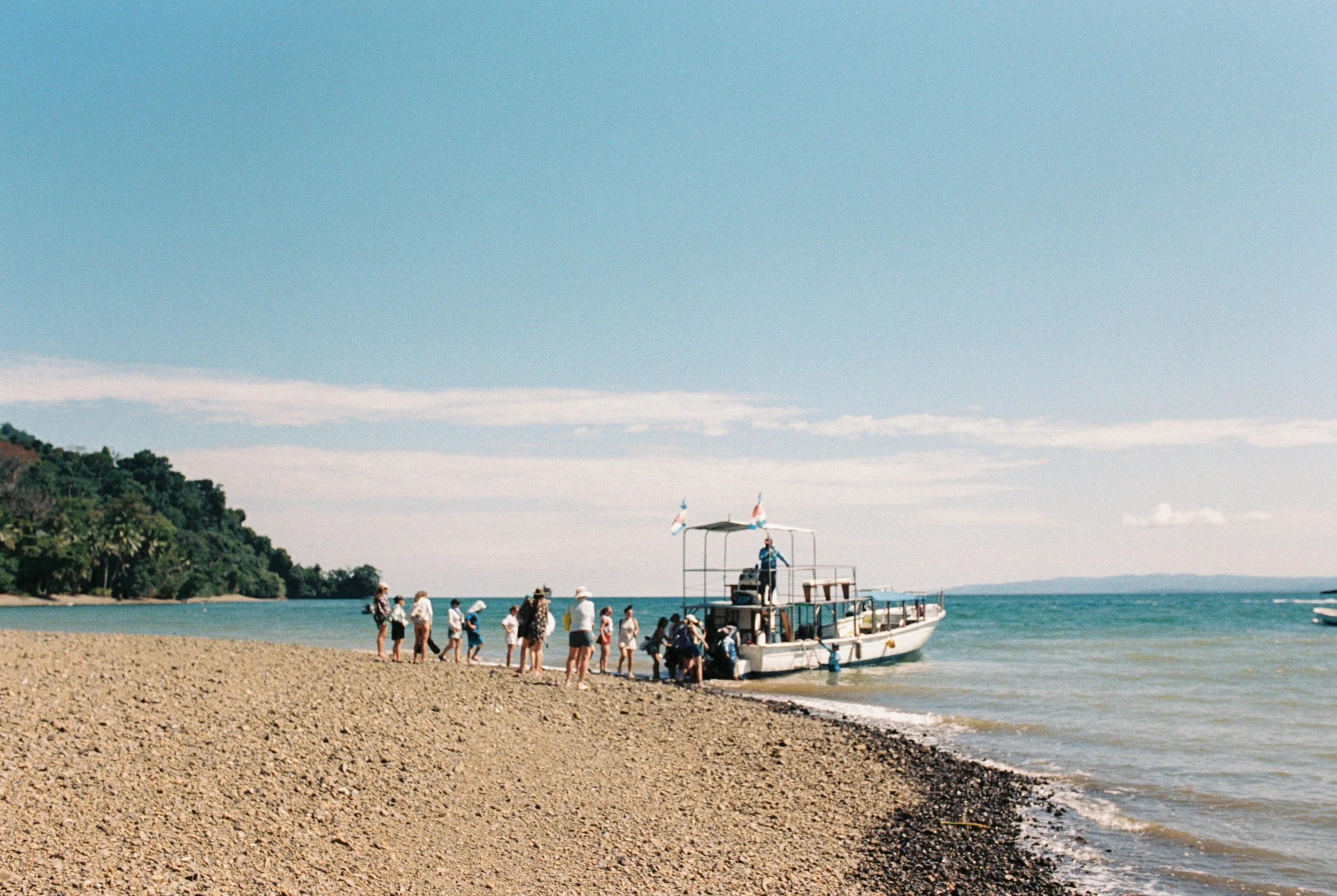 A boat docked at the shoreline with a group of people boarding or disembarking. The scene is by a lake or ocean, with trees on the hillside in the background and a clear sky overhead.
