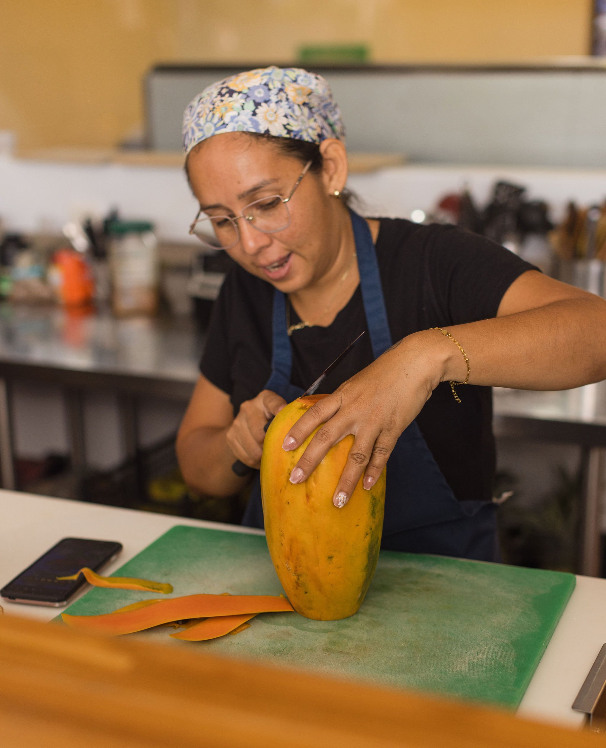 A woman with glasses and a floral cap is peeling a large yellow and green mango on a green cutting board, with a few mango peels on the table and a smartphone nearby.